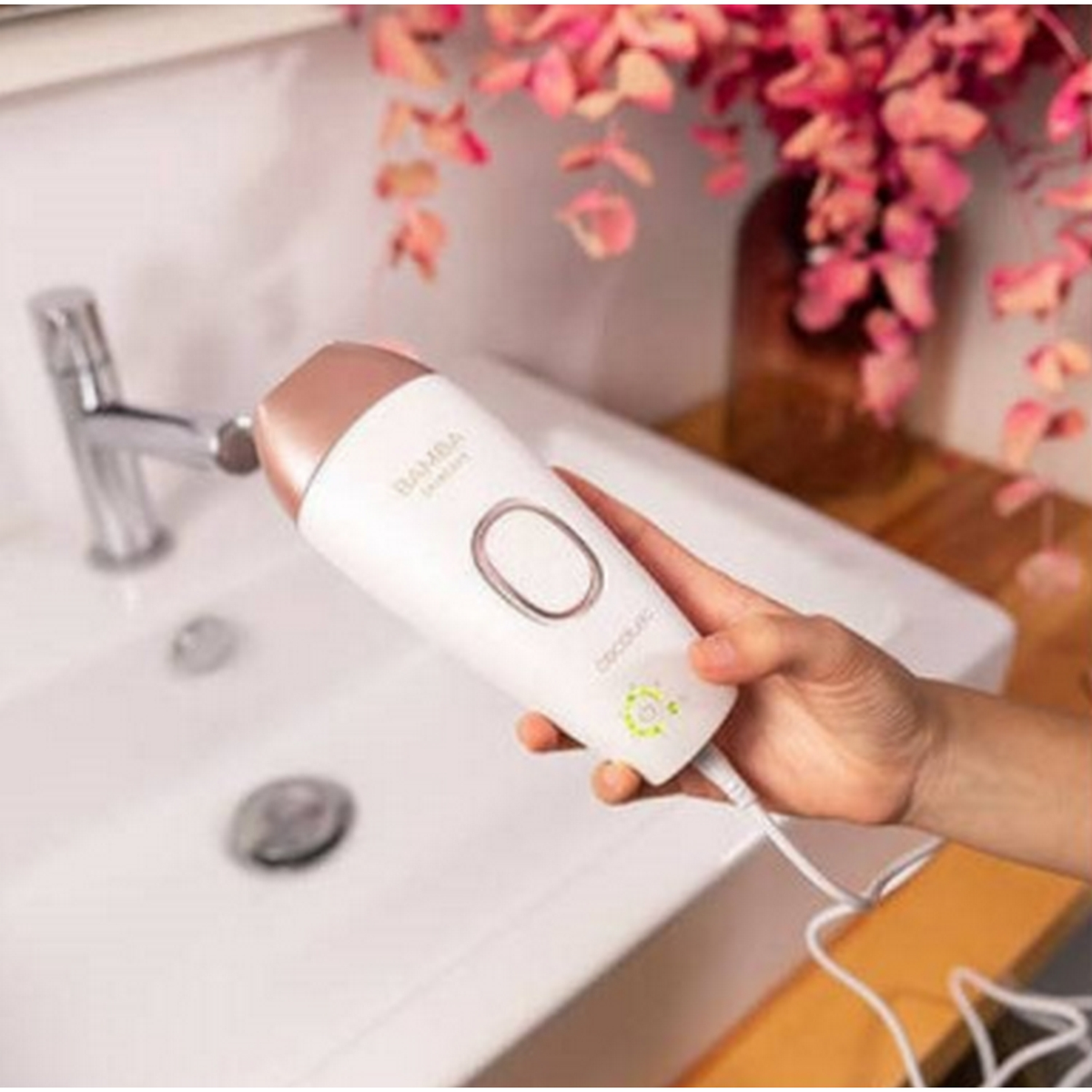 Białe i różowo-złote urządzenie do usuwania włosów, trzymane w dłoni. Umywalka i kwiaty w tle.