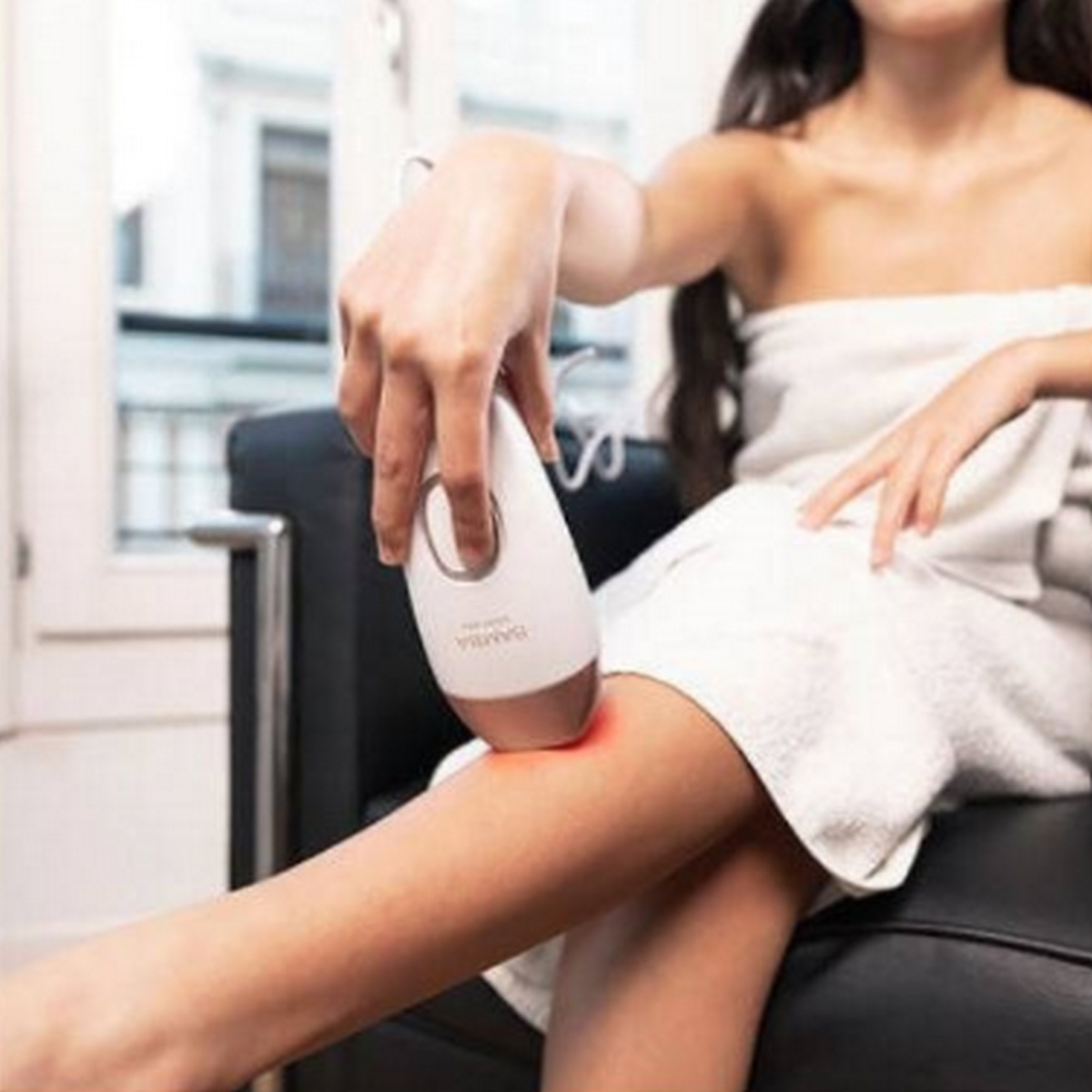 Kobieta używa urządzenia do usuwania włosów na nodze, siedząc na krześle owiniętym ręcznikiem.