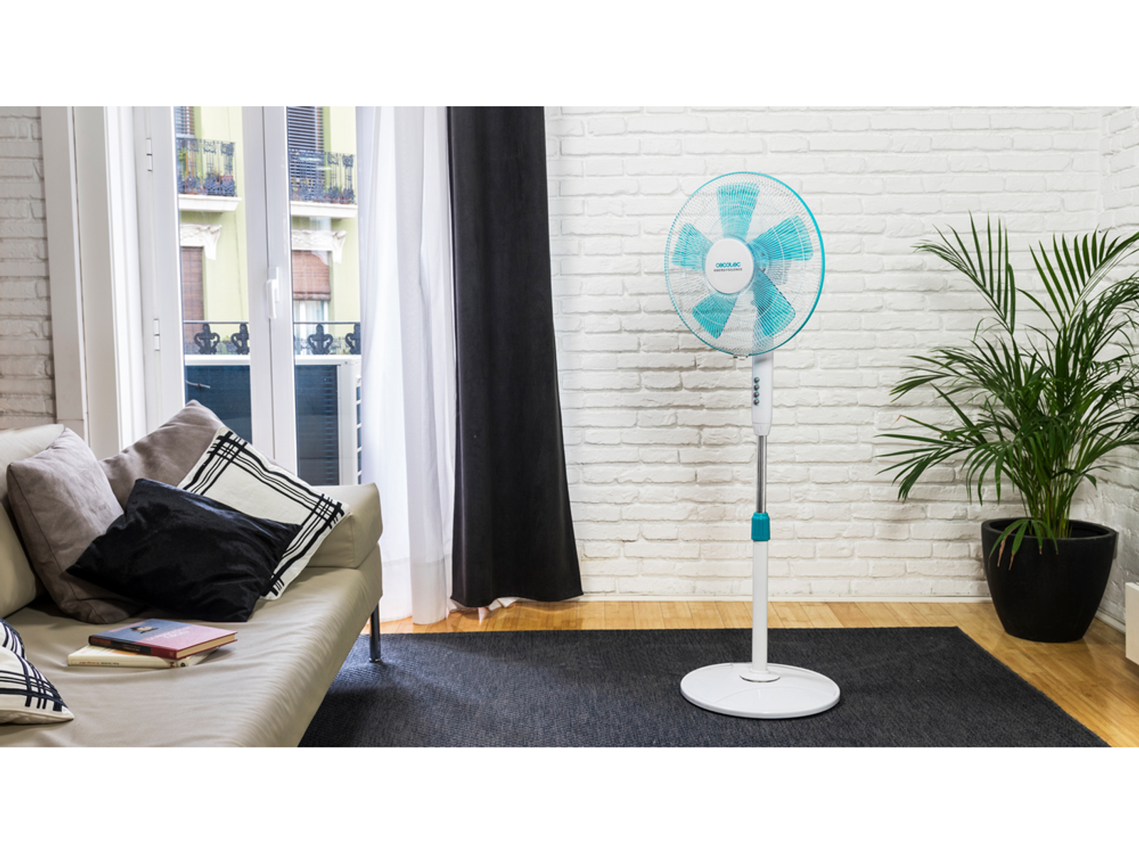 Biało-turkusowy wentylator stojący w pokoju z sofą i ceglaną ścianą.