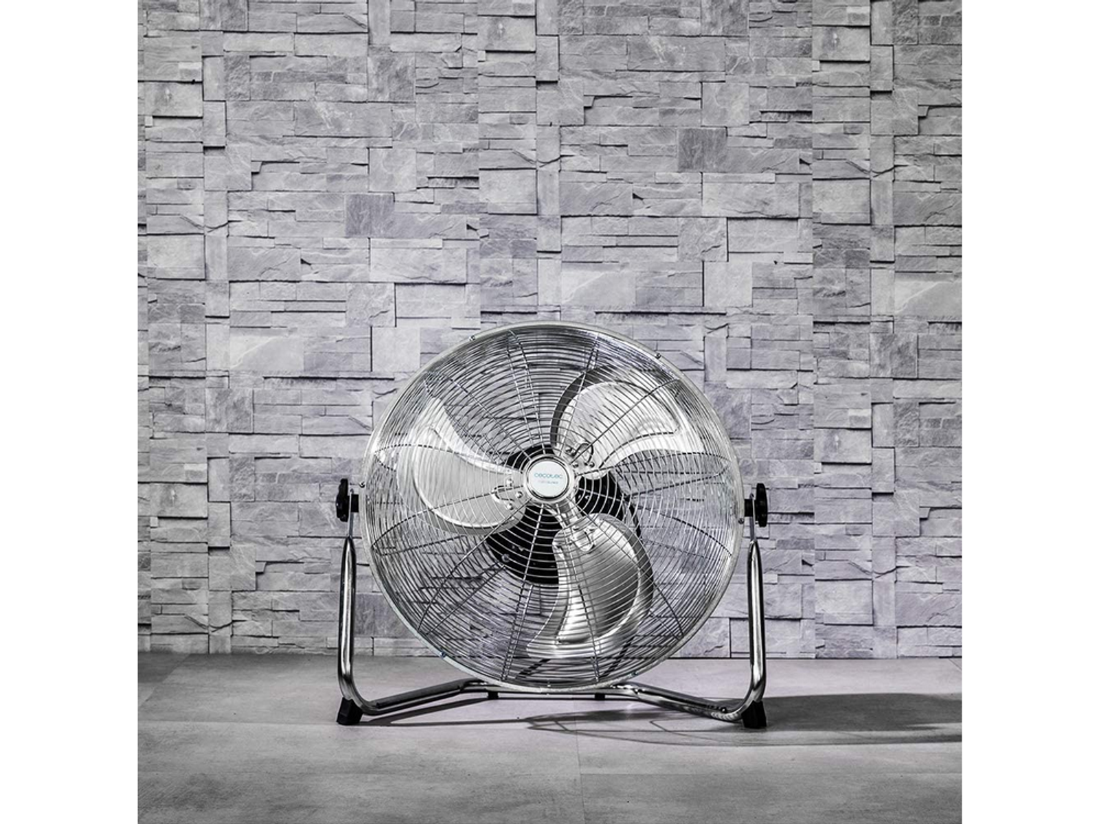 Duży srebrny wentylator z chromowaną podstawą, ustawiony przed szarą ścianą z kamiennych cegieł.