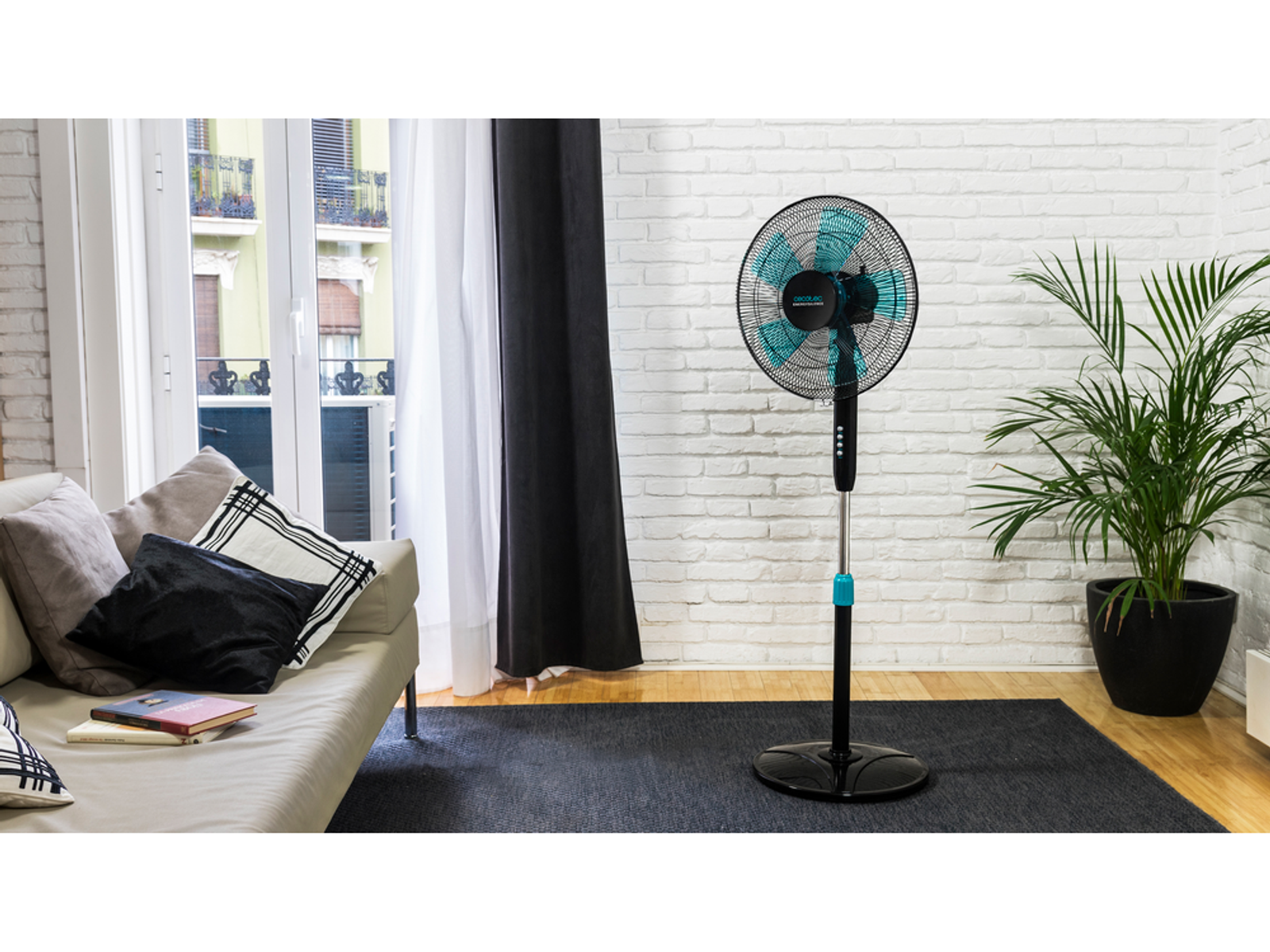 Czarno-turkusowy wentylator stojący w pokoju z sofą, książkami i roślinami, w pobliżu okna.