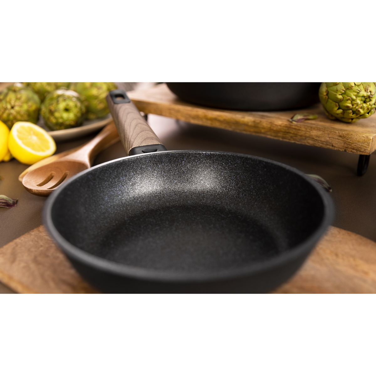Padella nera con manico in legno, circondata da carciofi e limoni.
