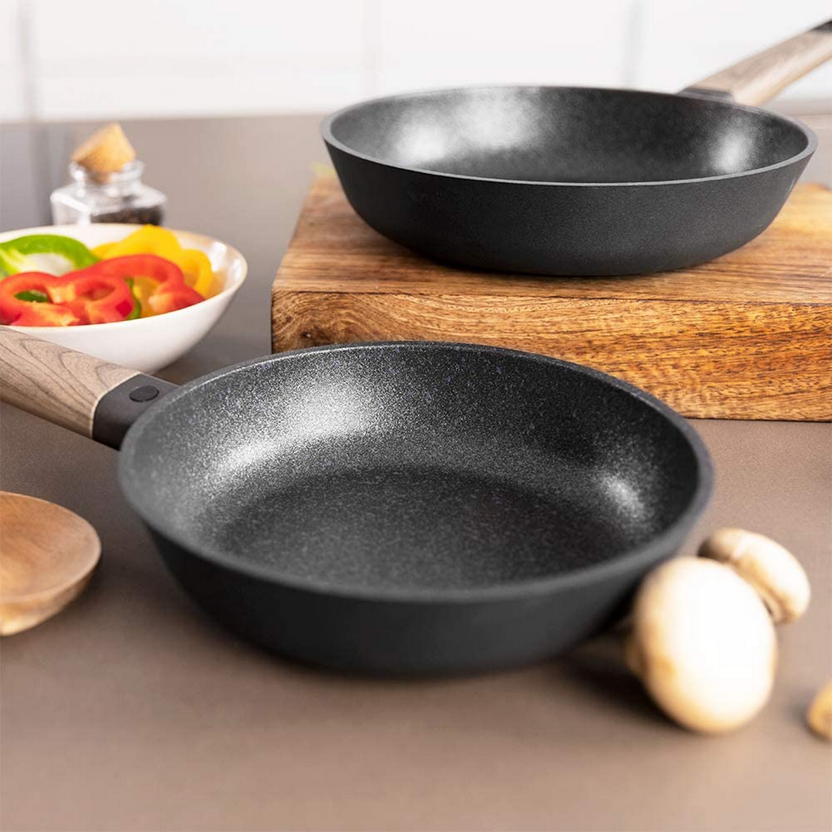 Due padelle nere su un tagliere di legno con una ciotola di verdure e una bottiglia di spezie.