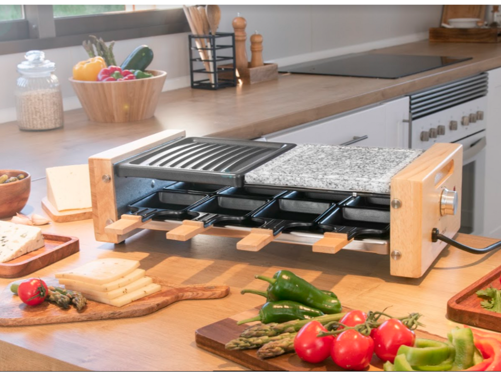 Grill do raclette z różnymi składnikami na drewnianym stole w kuchni.