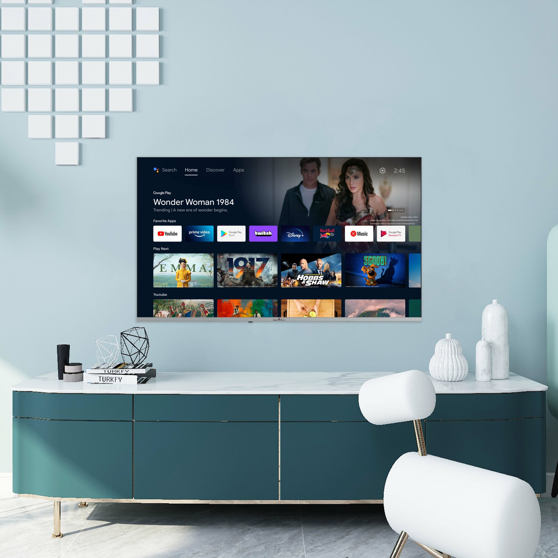 Ekran telewizora wyświetlający 'Wonder Woman 1984'. Telewizor nad szafką. Jasnoniebieskie tło.