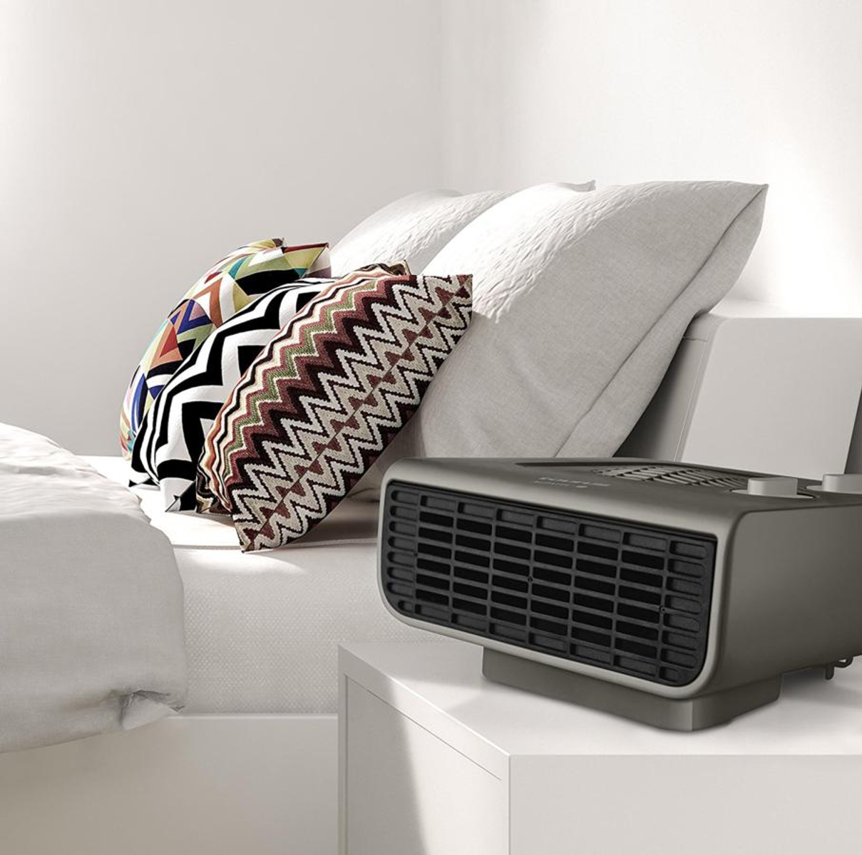 Szary grzejnik elektryczny na białej szafce nocnej obok łóżka z poduszkami.