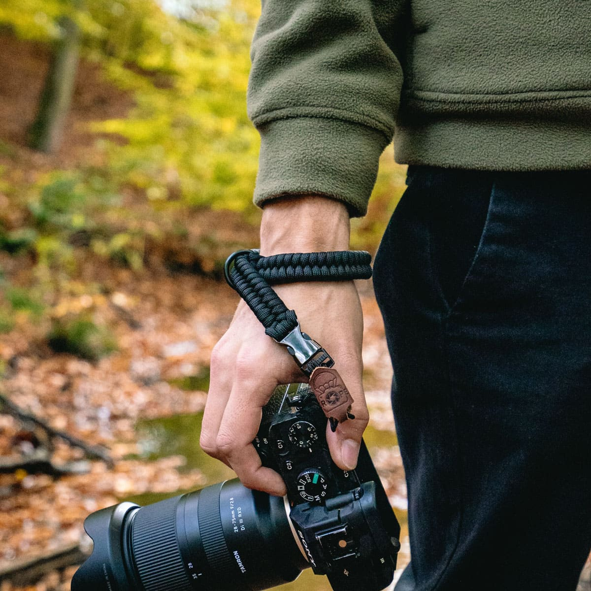 Una mano tiene una fotocamera con un cinturino nero, in un ambiente forestale.