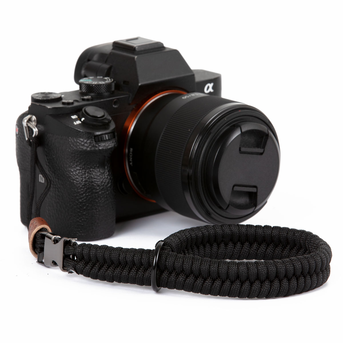 Fotocamera nera con obiettivo e cinturino da polso nero. Dettaglio in pelle marrone. Sfondo bianco.