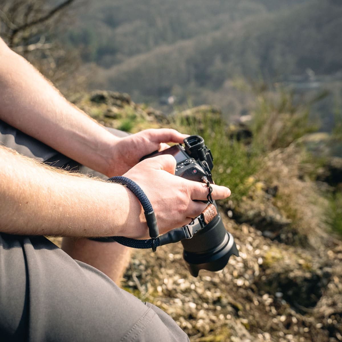 Person hält Kamera mit Handschlaufe, draußen mit unscharfem Hintergrund der Natur.