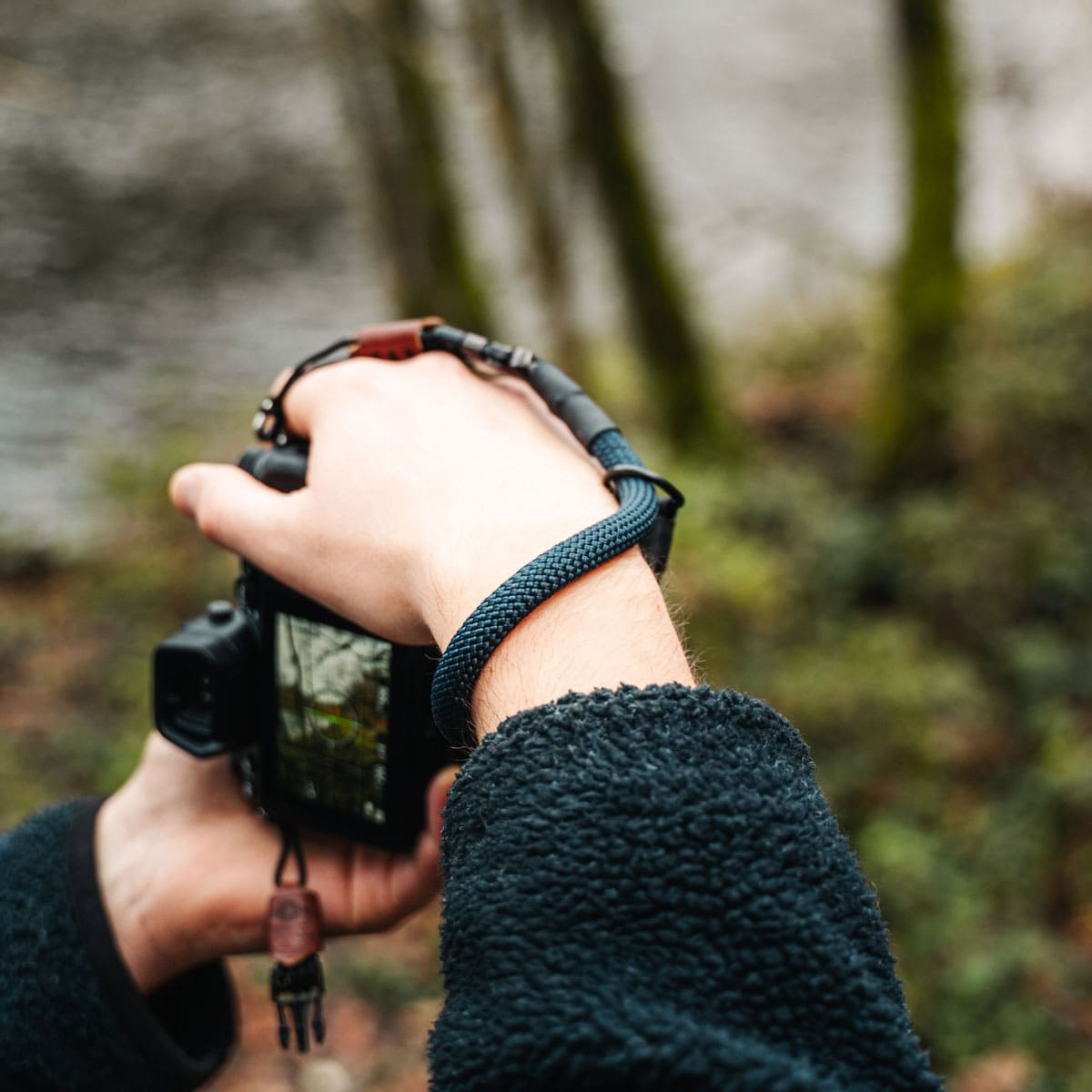 Person hält eine Kamera mit blauem Gurt. Der Hintergrund zeigt verschwommenen Wald und Wasser.