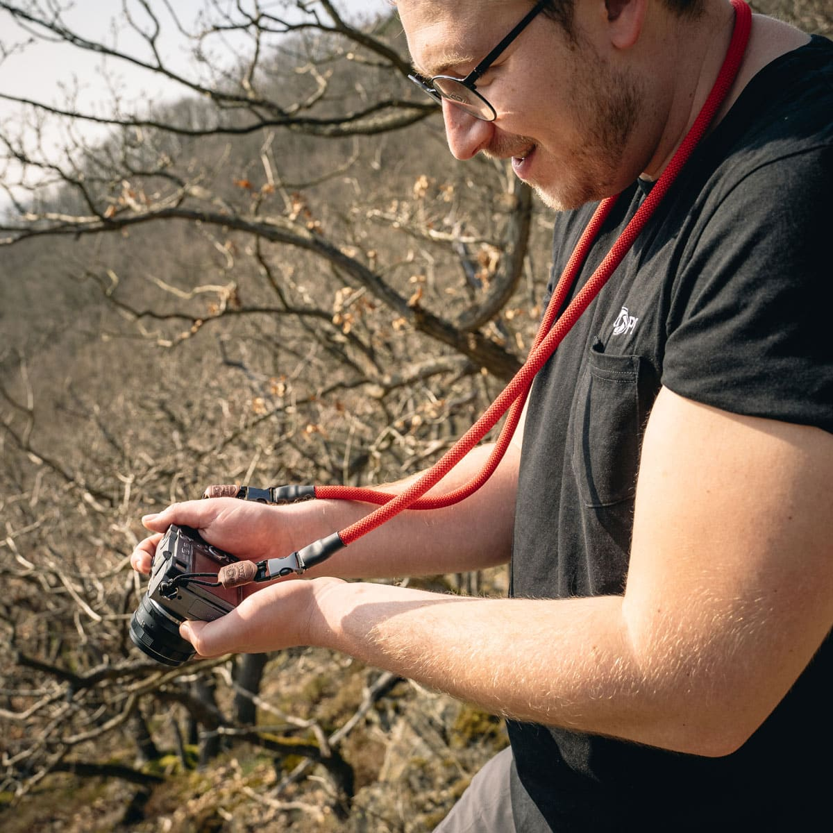 Ein Mann hält eine Kamera mit rotem Kameragurt, lächelnd draußen in der Natur.