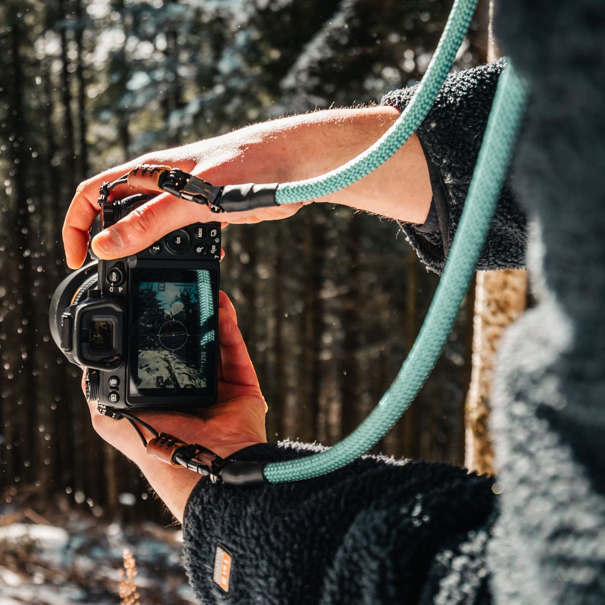 Eine Person, die eine Kamera mit blauem Gurt in einem verschneiten Wald hält, Kamerabildschirm sichtbar.