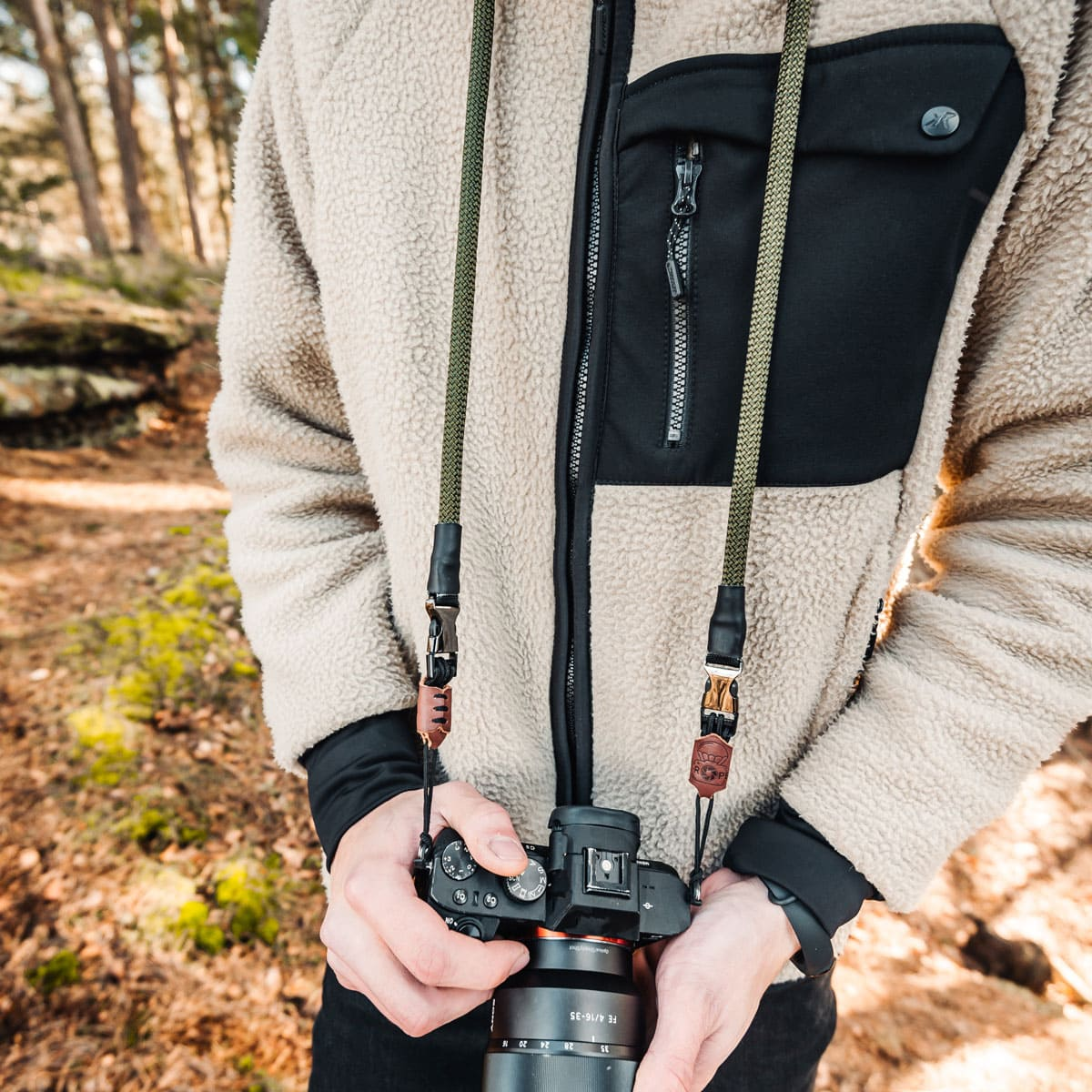 Eine Person mit einer Fleecejacke, die eine Kamera mit Gurt in einem Wald hält.