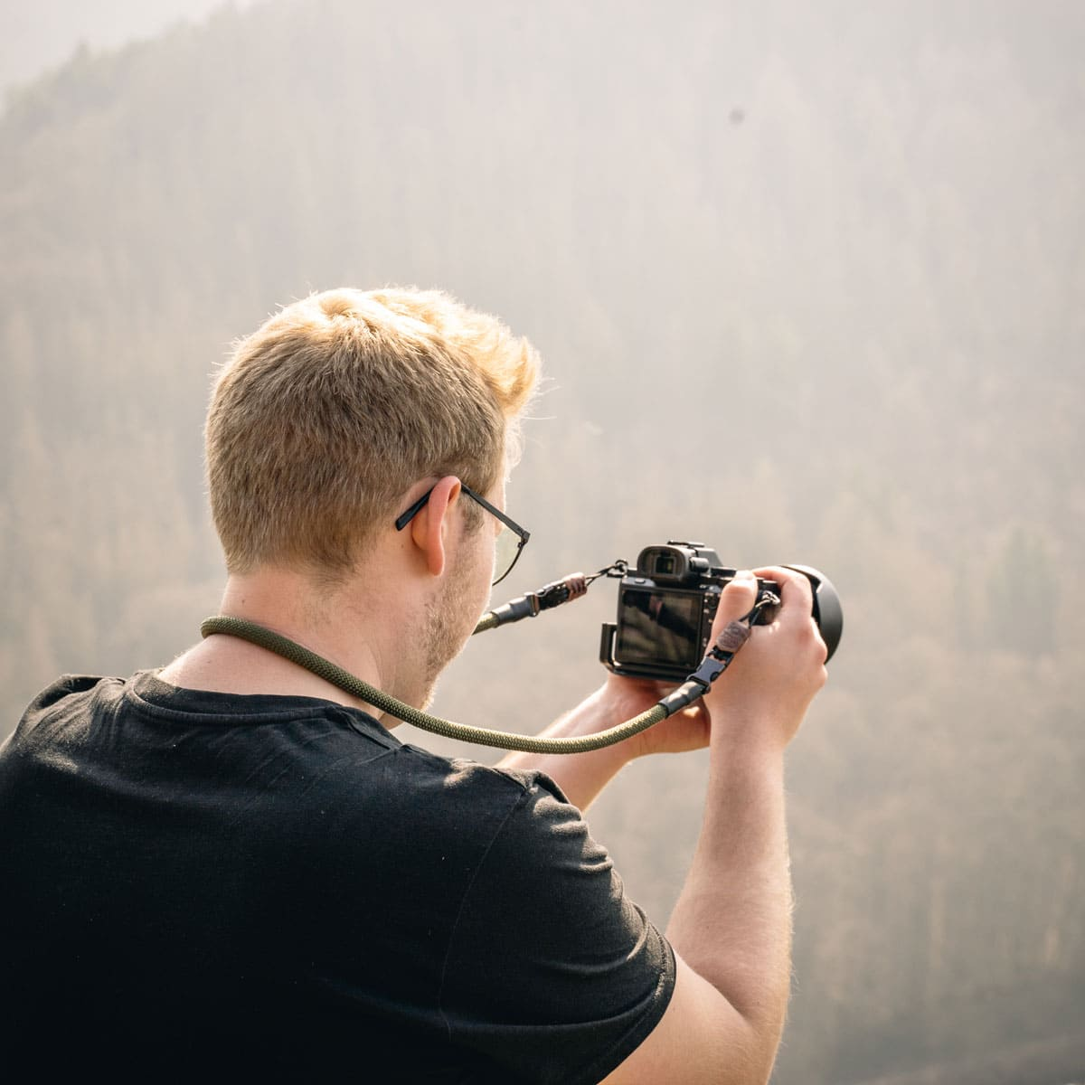 Mann fotografiert mit einer Kamera in natürlicher Umgebung, trägt Brille und schwarzes Hemd.