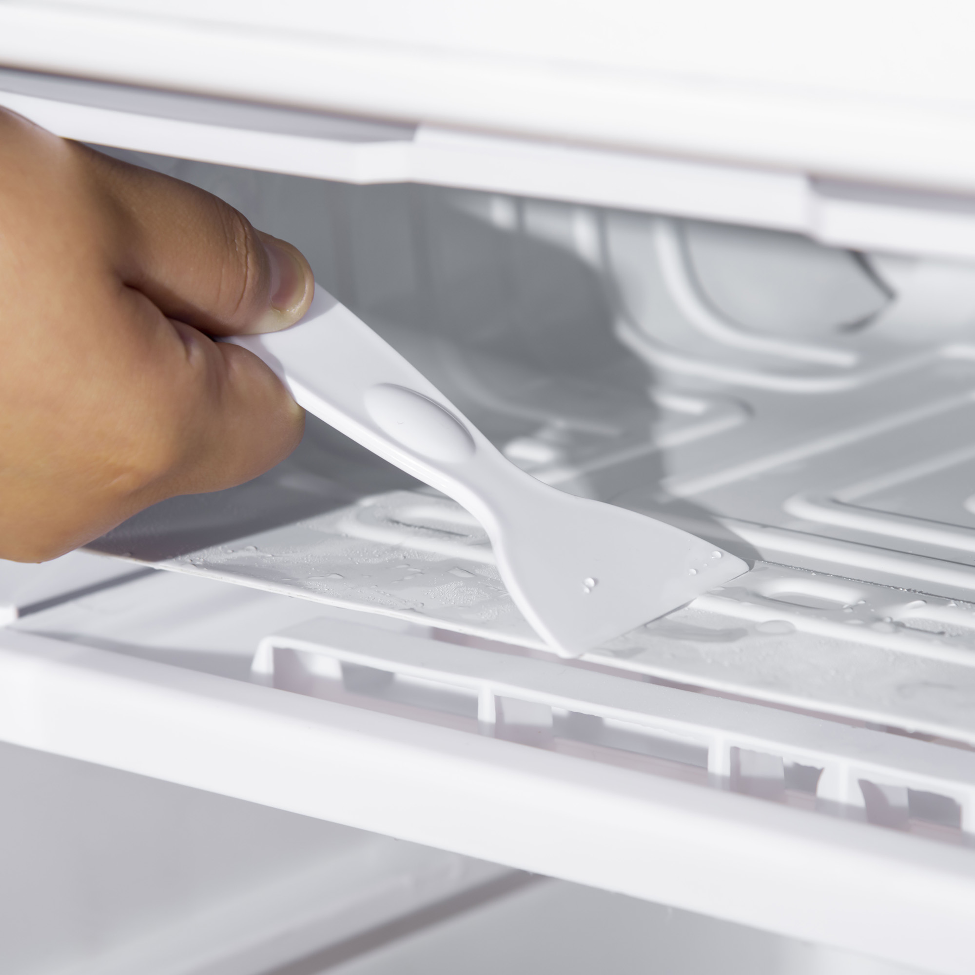 Una mano usando un raspador de plástico blanco para quitar el hielo del interior de un refrigerador.