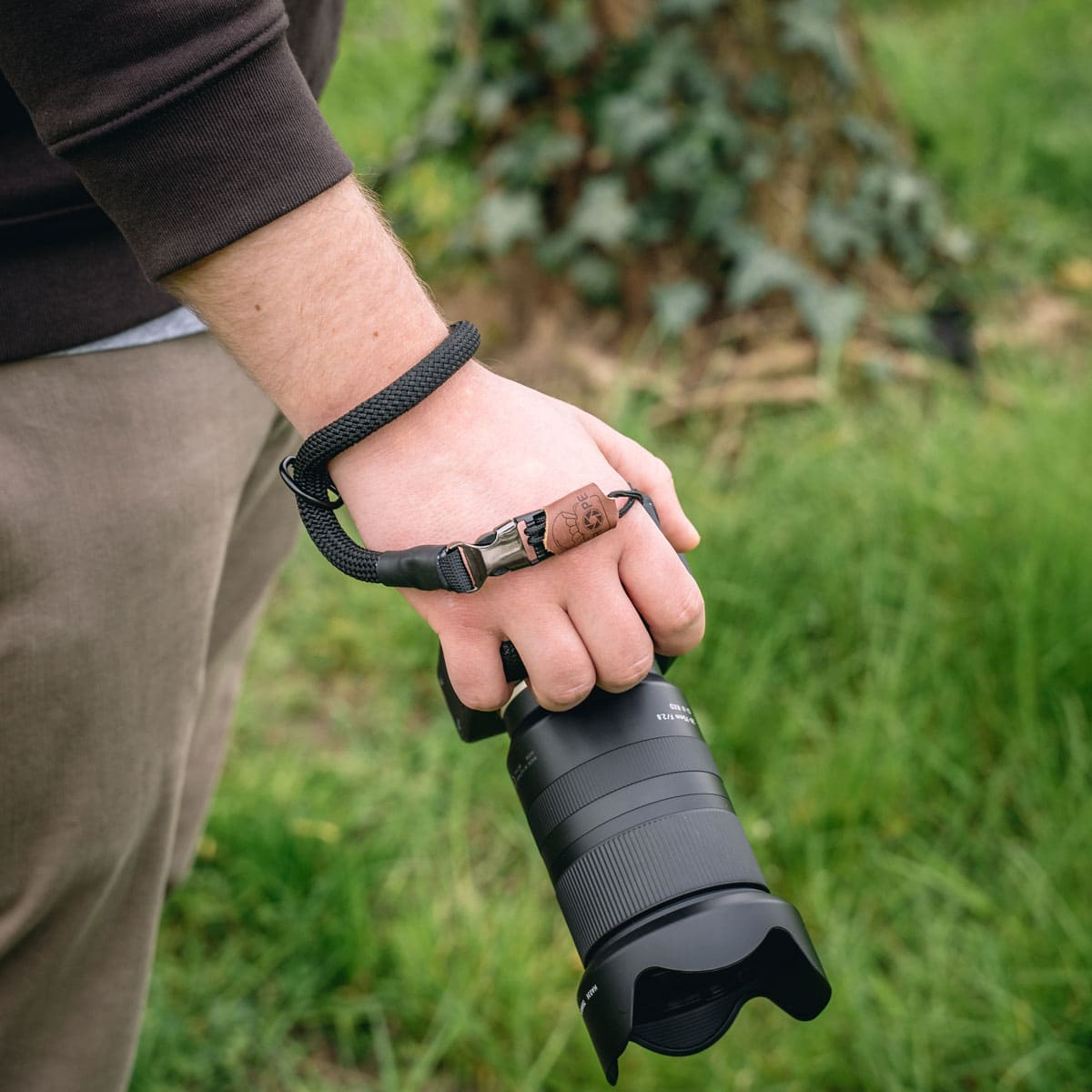 Mann hält Kamera mit Handschlaufe in grünem Grasbereich mit Baum.