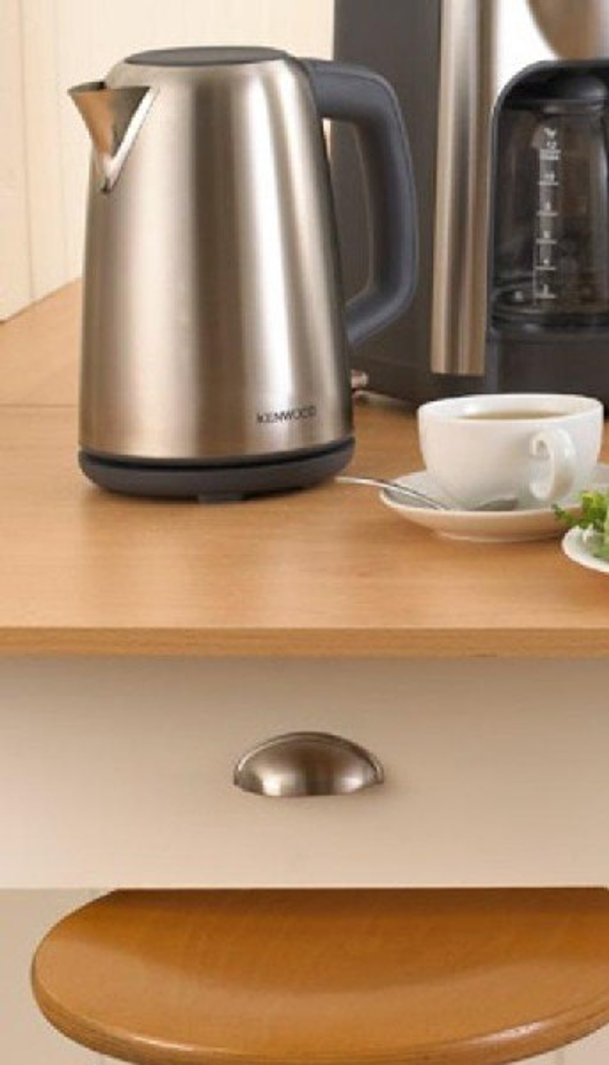 Czajnik ze stali nierdzewnej i filiżanka herbaty na drewnianej powierzchni z uchwytem szuflady poniżej.