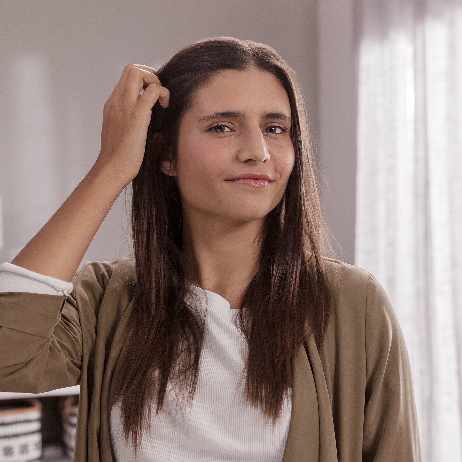 Una giovane donna con lunghi capelli castani che si tocca i capelli, guarda la fotocamera con un leggero sorriso.