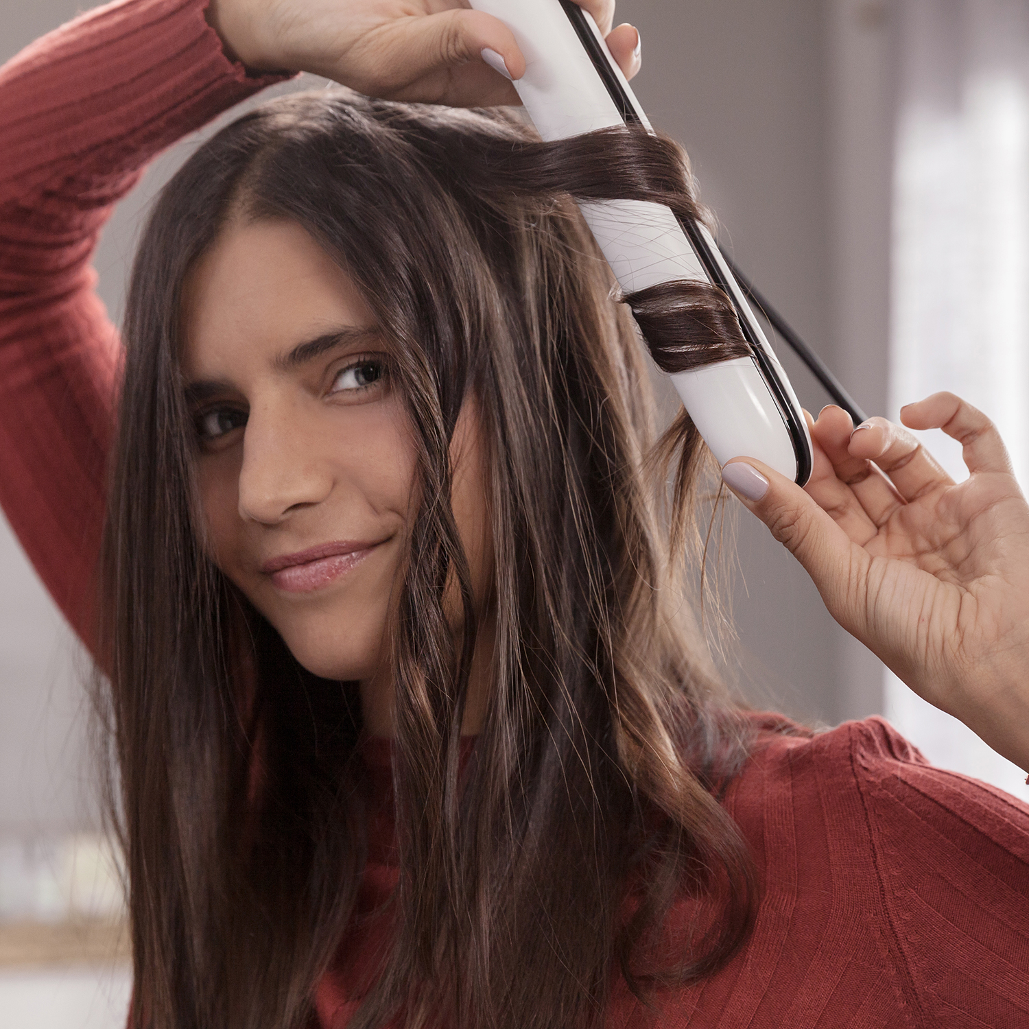 Una donna che si arriccia i capelli con un arricciacapelli bianco e nero. Indossa un maglione rosso e sorride.
