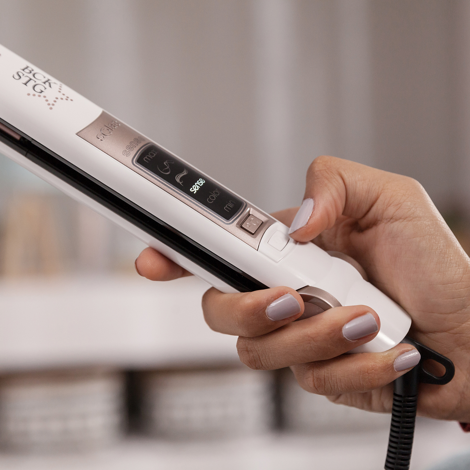 Una mano tiene una piastra per capelli bianca con dettagli oro rosa. La piastra ha un display digitale e pulsanti di controllo.