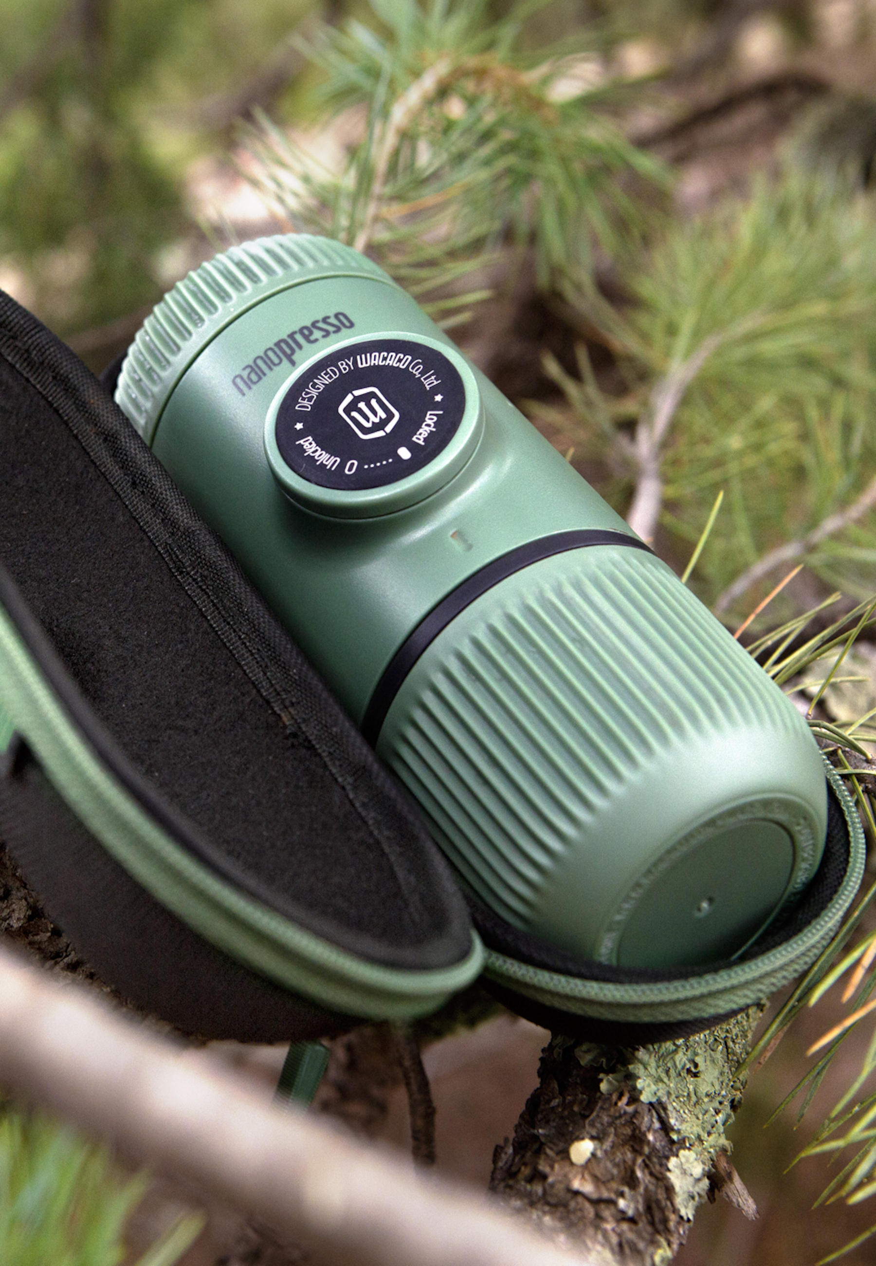 Macchina per espresso portatile verde nella sua custodia su un ramo, circondata dalla natura. L'etichetta dice 'nanopresso'.
