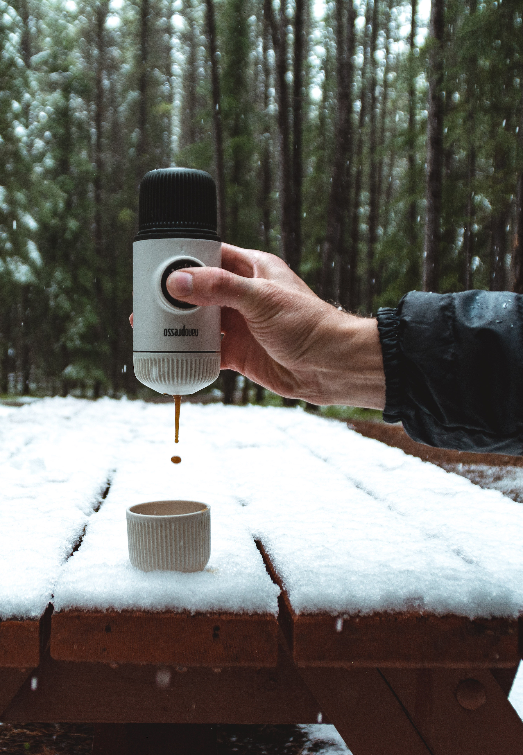 Eine Person, die Kaffee aus einer weißen, tragbaren Espressomaschine in eine Tasse auf einem schneebedeckten Tisch gießt.