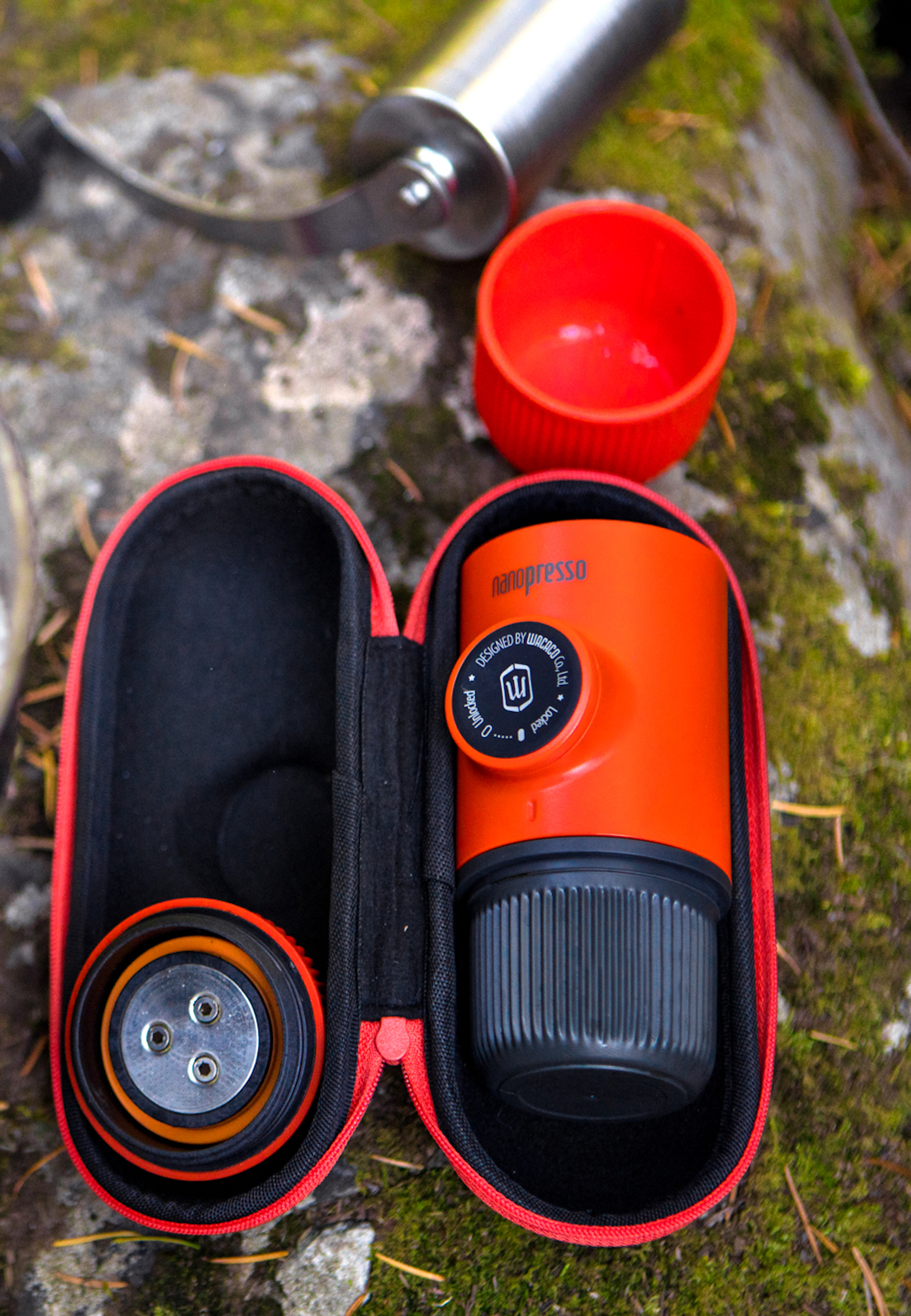Orange tragbare Espressomaschine mit schwarzem Gehäuse, Mühle und roter Kappe auf einem Stein.