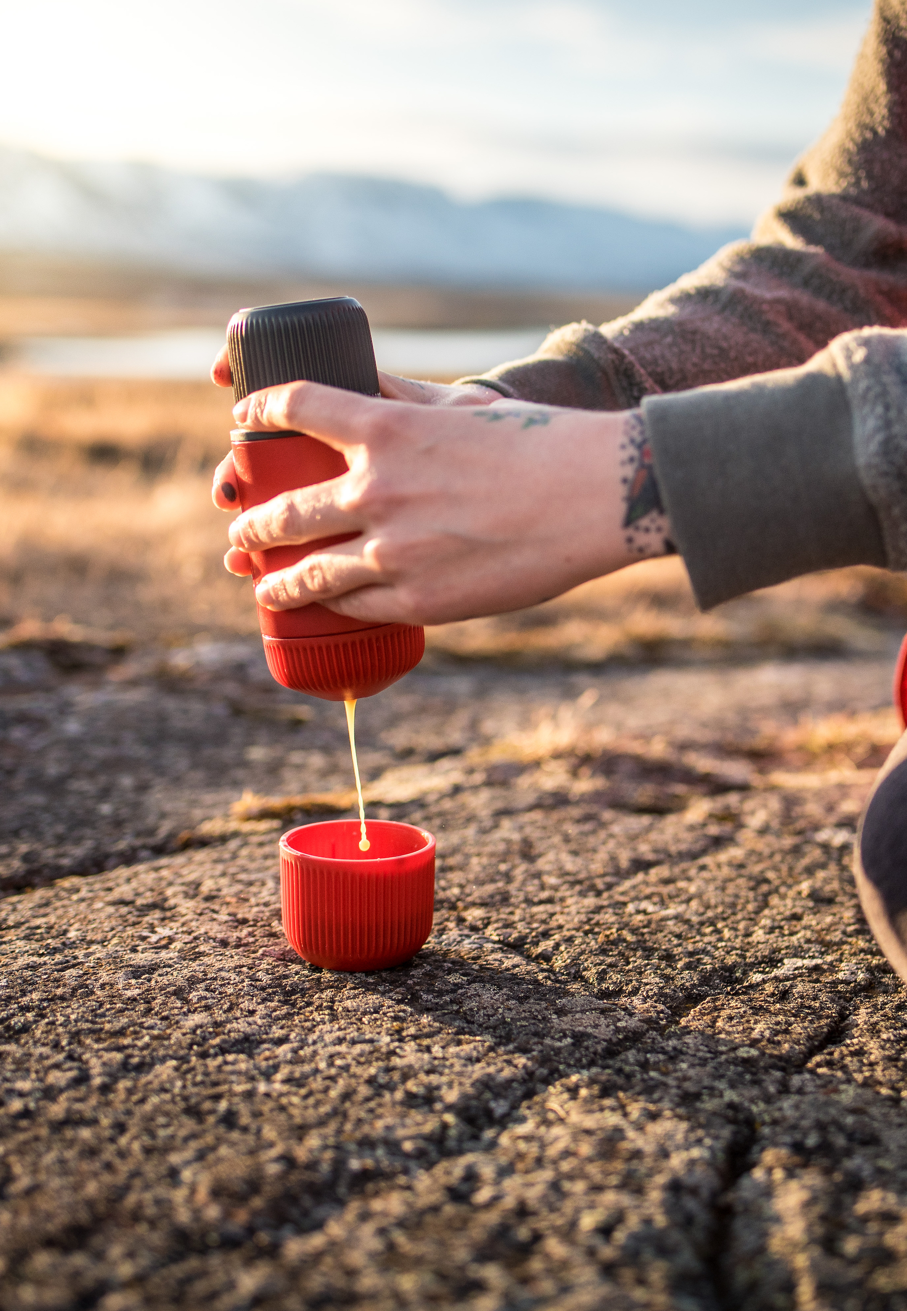Eine Person gießt Flüssigkeit aus einem rot-schwarzen Behälter in eine rote Tasse, draußen mit Bergblick.