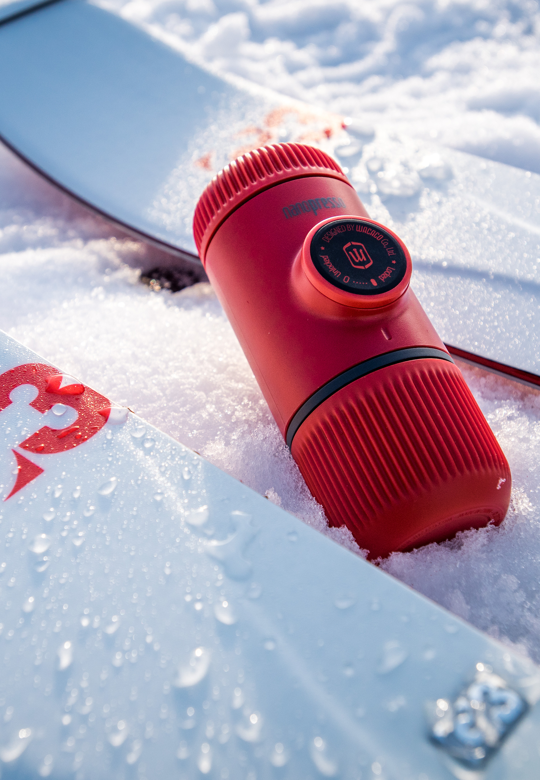 Roter tragbarer Espressomacher im Schnee, zwischen Skiern. Schwarzer Ring.