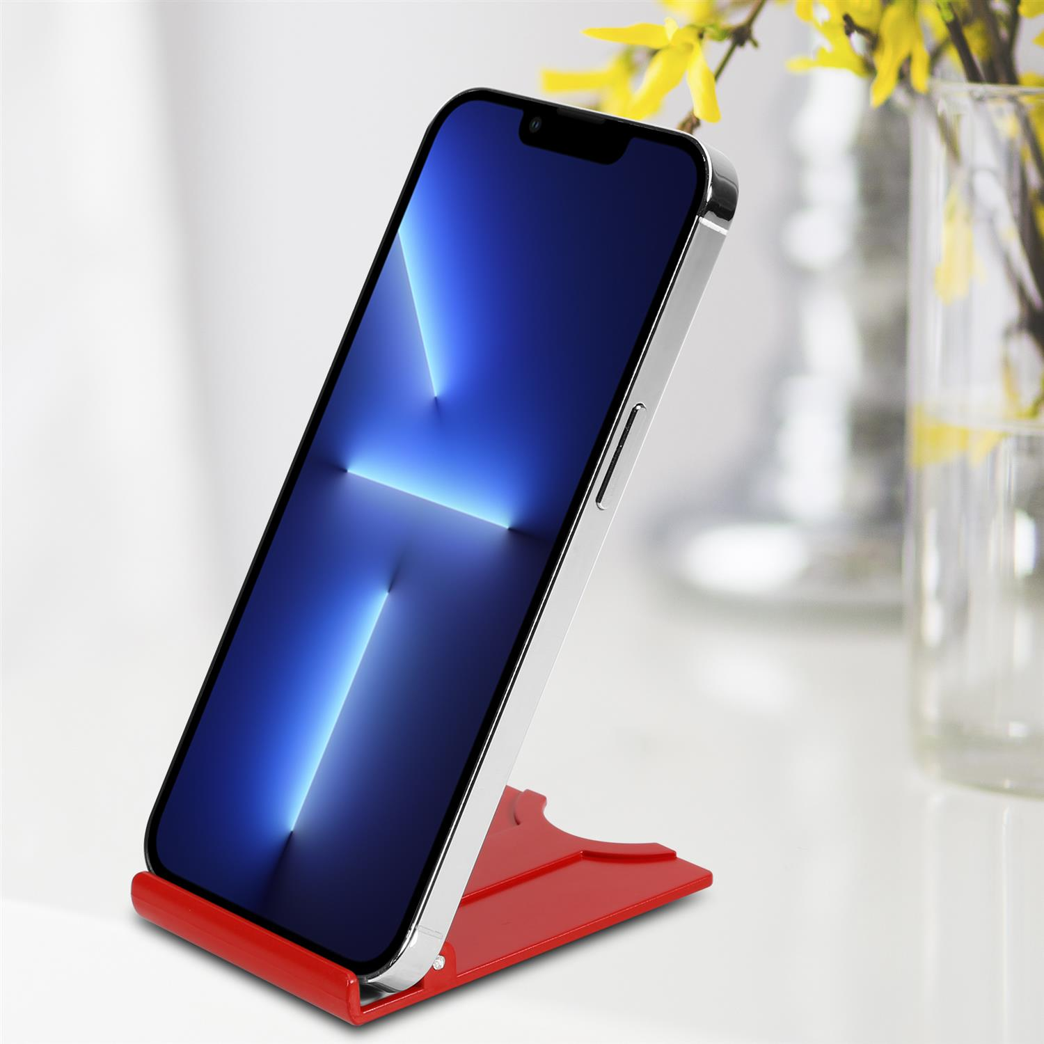 Ein Telefon auf einem roten Ständer, abgewinkelt, mit blauem Bildschirm. Hintergrund zeigt weiße Oberfläche und Vase.