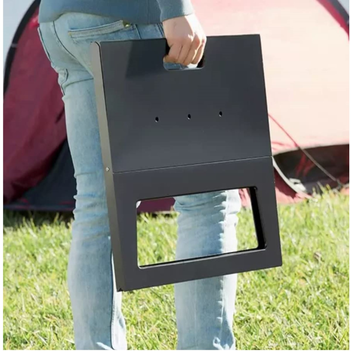 Eine Person hält einen schwarzen Klappgrill. Die Person steht auf dem Gras.