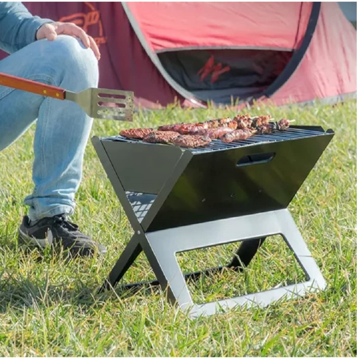 Eine Person grillt Essen auf einem tragbaren schwarzen Grill auf Gras, im Hintergrund ein rotes Zelt.
