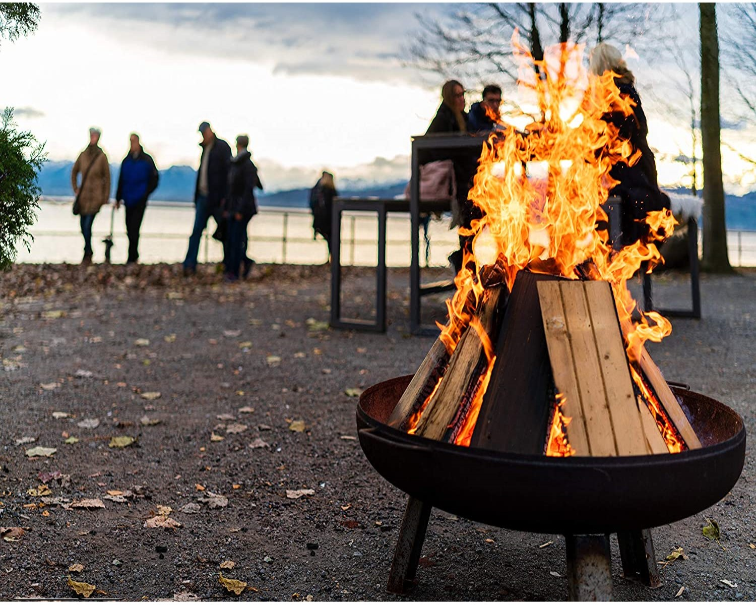 Ein Lagerfeuer brennt in einer Feuerstelle aus Metall. Menschen stehen im Hintergrund in der Nähe von Wasser.