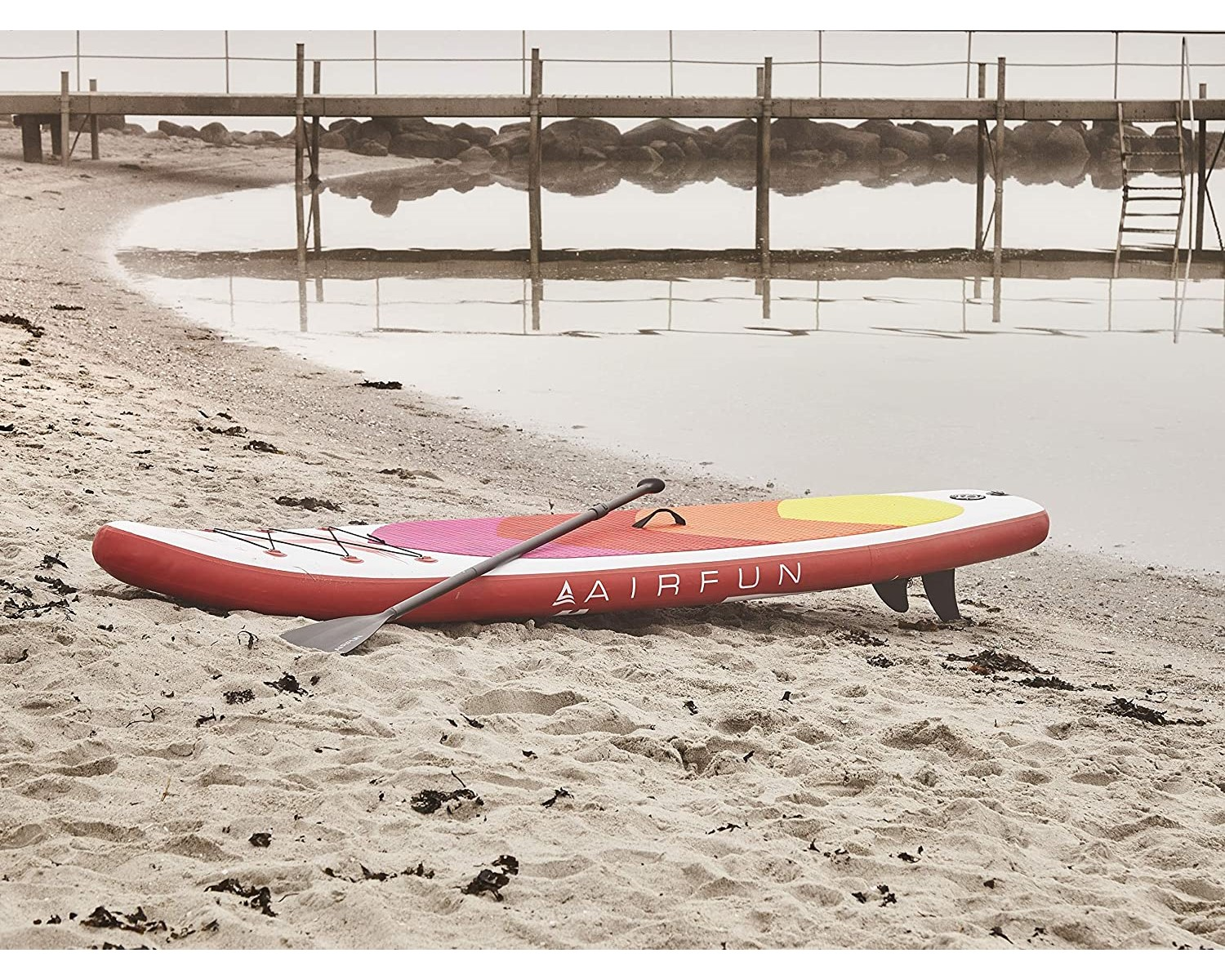 Rotes und weißes AIRFUN-Paddelboard an einem Sandstrand in Wassernähe. Ein Paddel liegt auf dem Board.