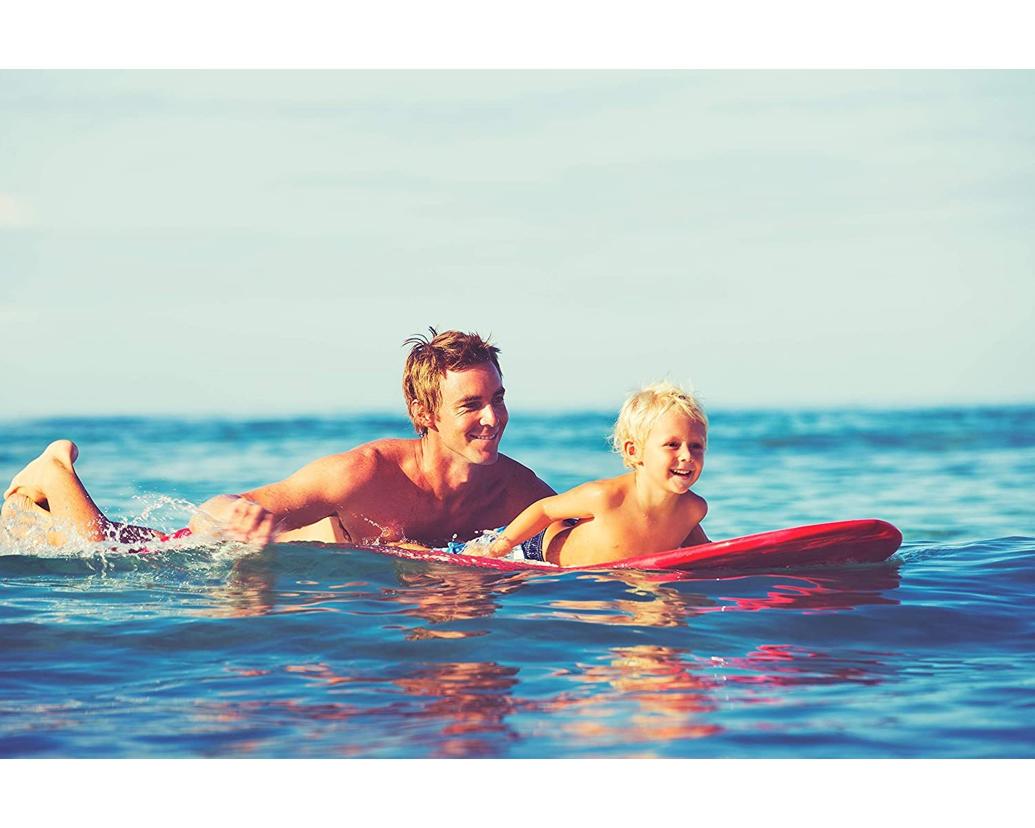 Vater und Kind surfen auf einem roten Brett im Meer. Blaues Wasser und Himmel.