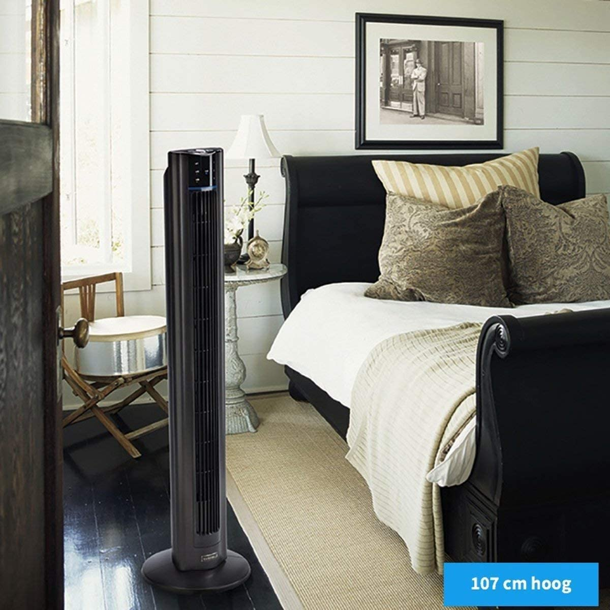 Schwarzer Turmventilator in einem Schlafzimmer. Ein Bett, Tisch und ein gerahmtes Bild sind ebenfalls sichtbar.