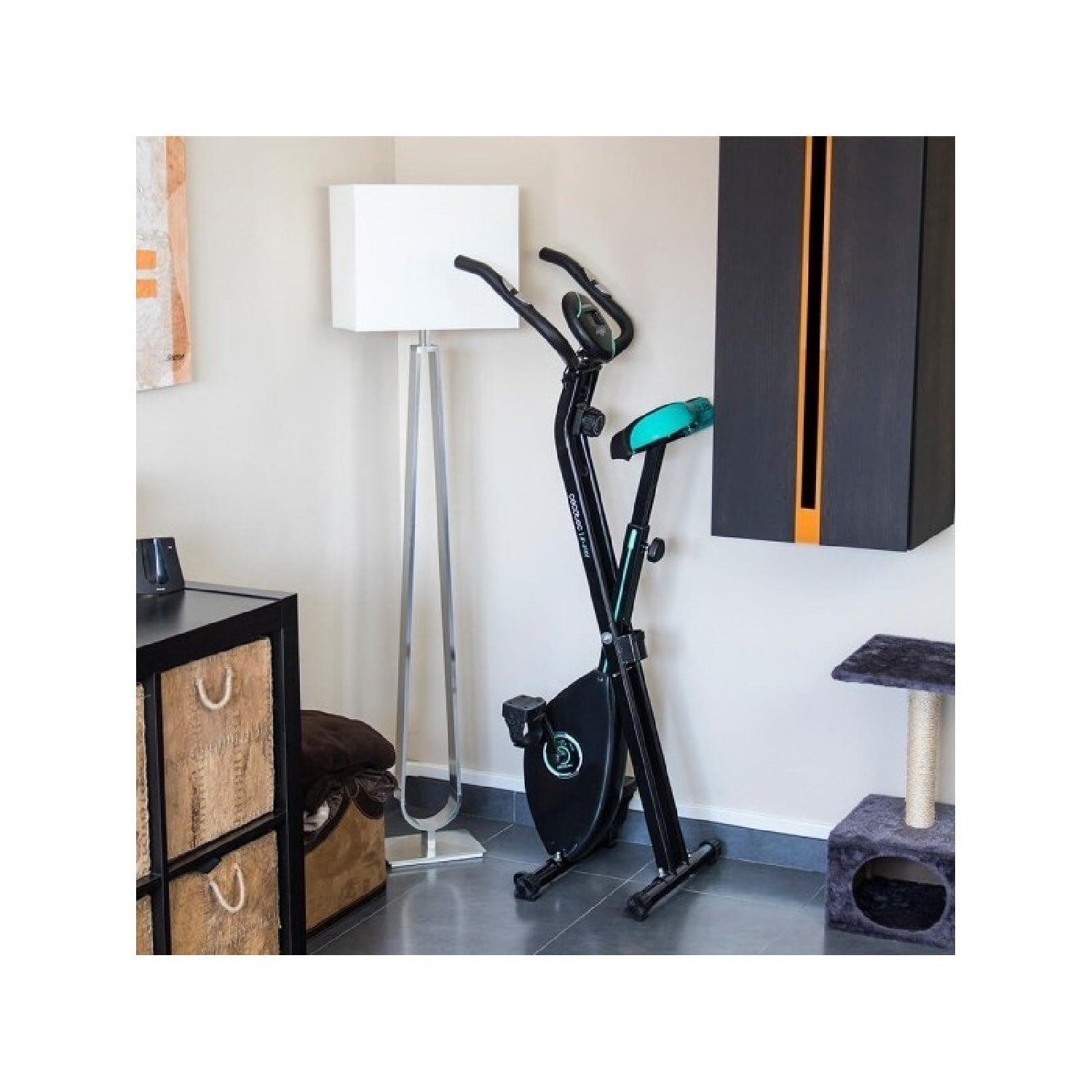 Una bicicleta estática en una habitación. Es negra y verde, cerca de una lámpara y un armario.