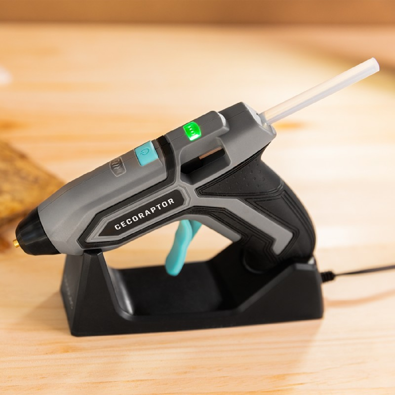 Szary pistolet do kleju na czarnej podstawie. Świeci się zielone światło. Napis 'CECORAPTOR'.