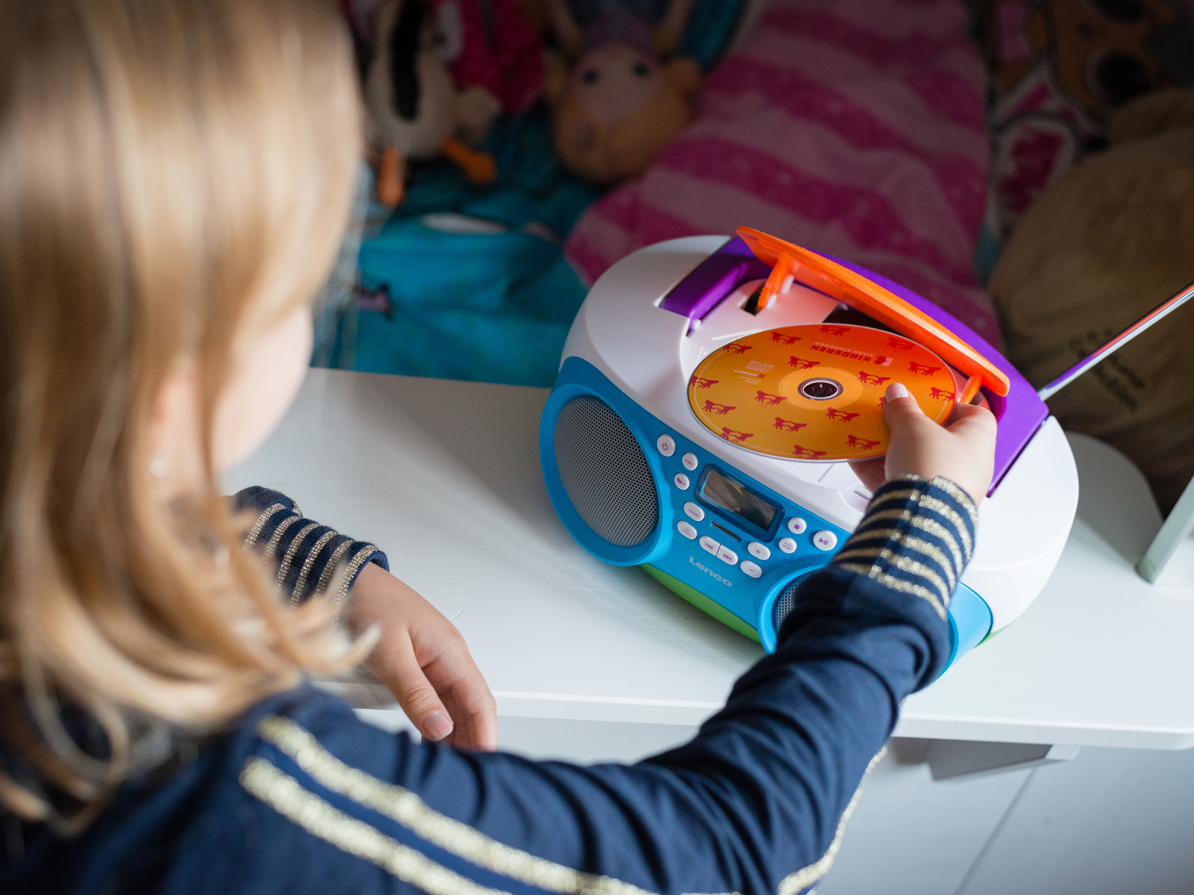Ein Kind legt eine CD in eine blau-weiße Boombox. Die Boombox ist weiß mit lila Akzenten.