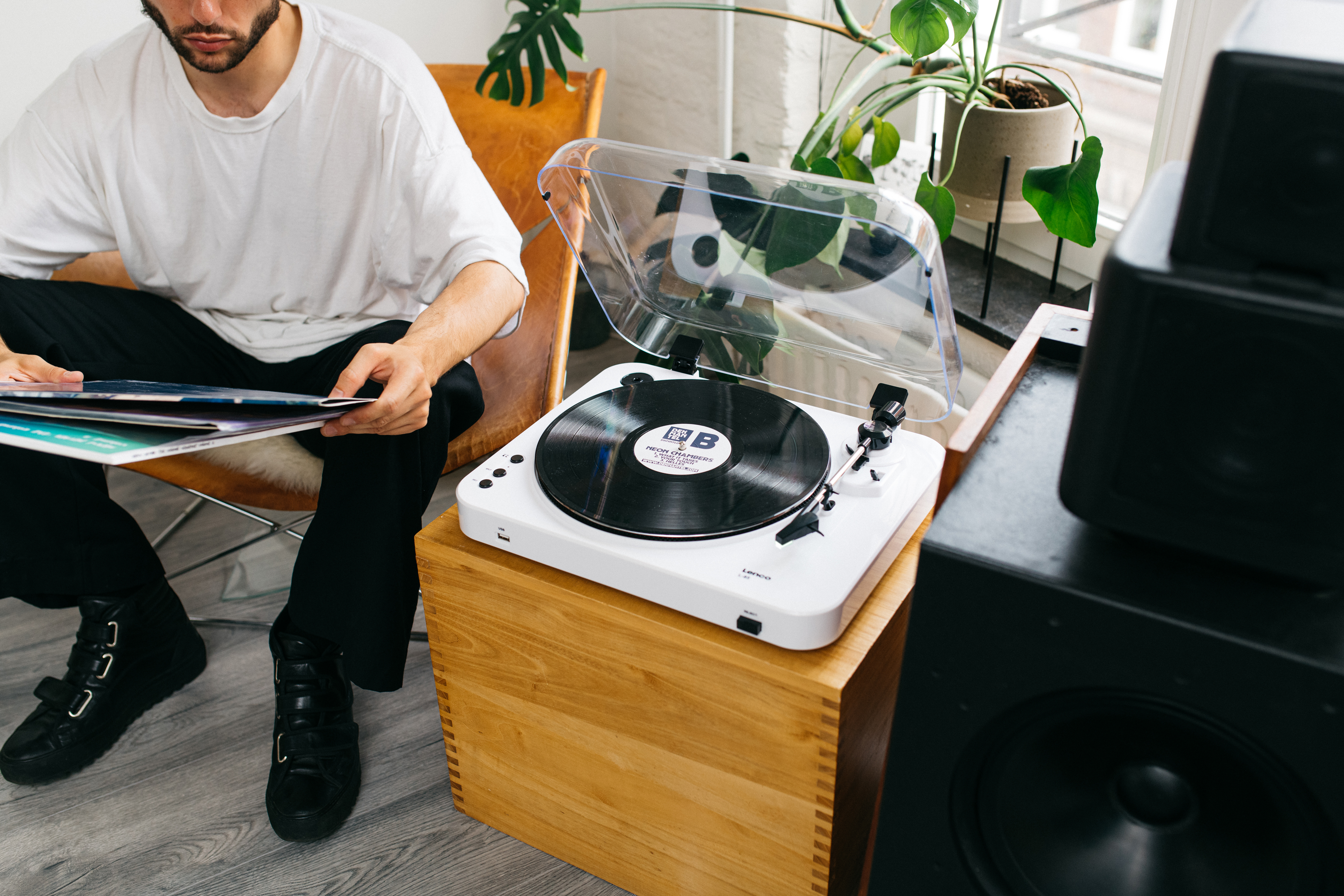 Mann sitzt, hält Schallplatten, in der Nähe eines weißen Plattenspielers auf einer Holzkiste. Lautsprecher rechts.