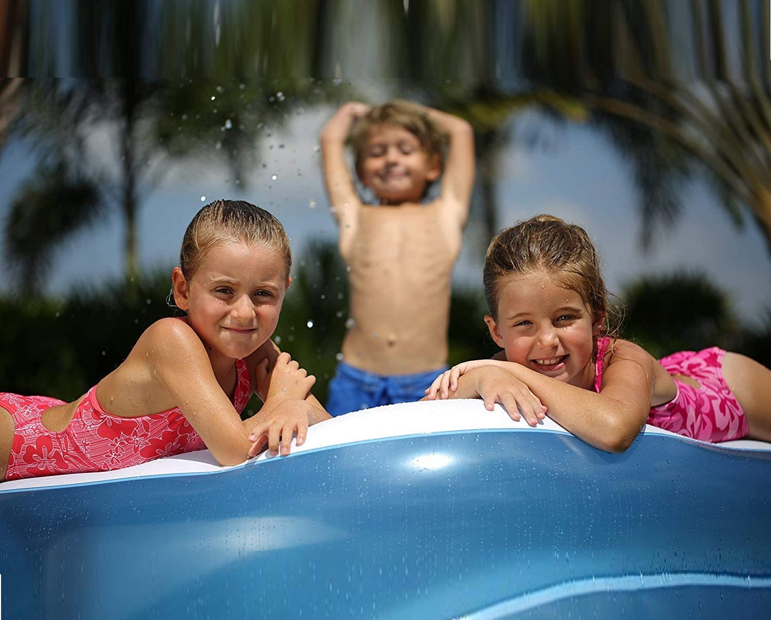 Drei Kinder in einem Pool, zwei Mädchen lächeln vorne, ein Junge hinten lächelt.