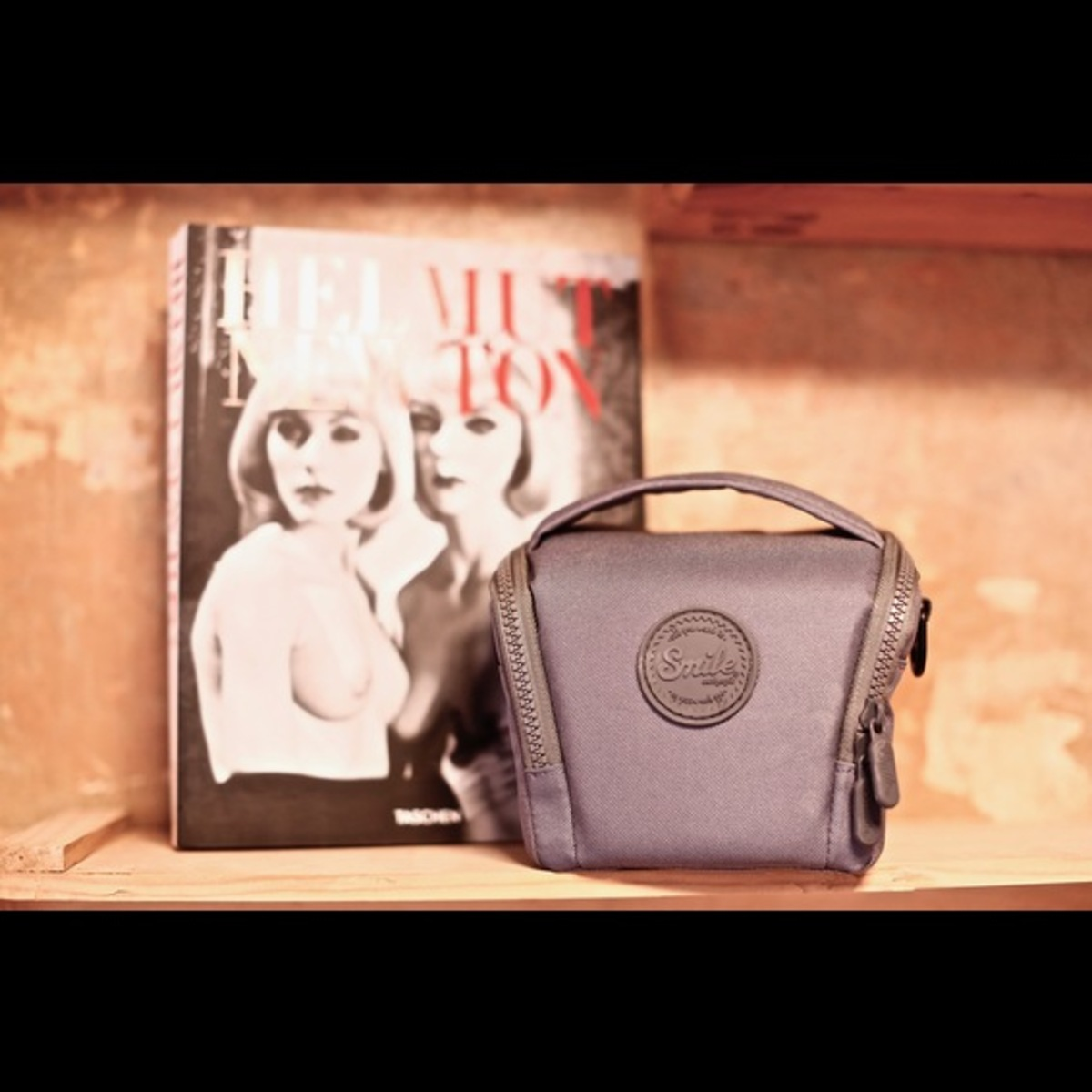 Pequeña bolsa de cámara gris en un estante. Un libro y una pared de madera en el fondo.