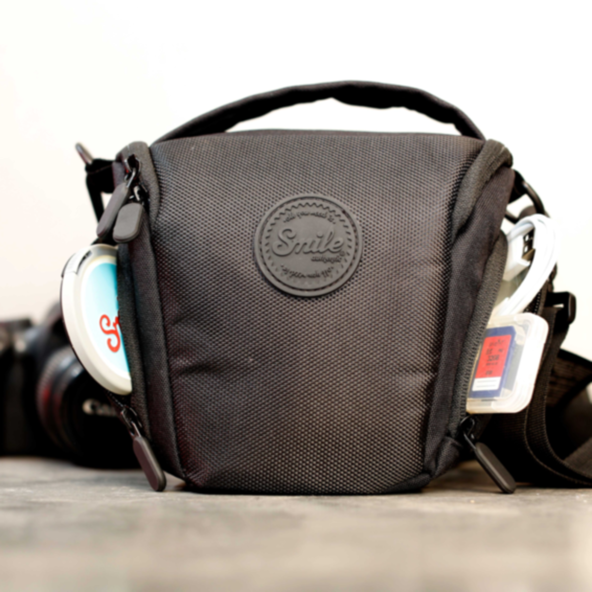 Bolsa de cámara negra con fondo blanco. Tiene logo Smile y accesorios en bolsillos.