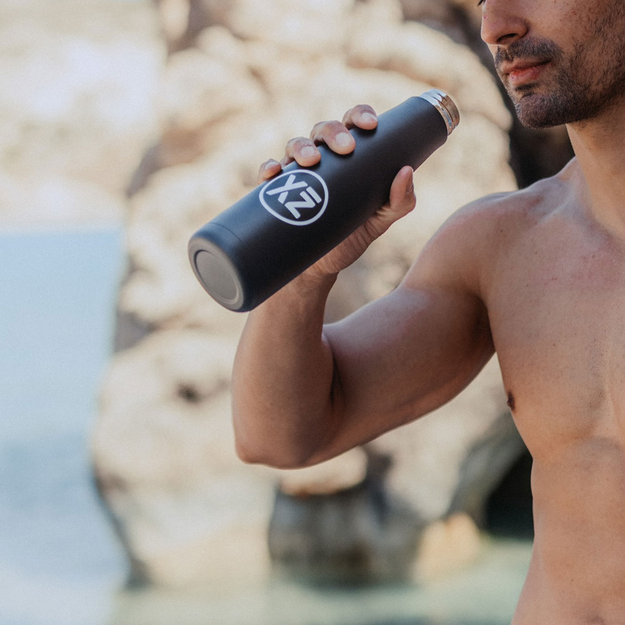 Hombre sosteniendo botella de agua negra con logo blanco XZ, cerca de la playa.