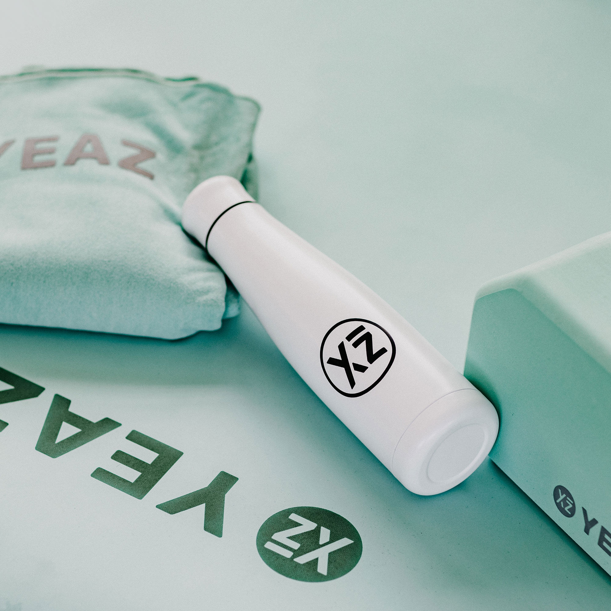 Una botella de agua blanca con un logo, una esterilla de yoga y un bloque verde menta. El fondo es verde claro.