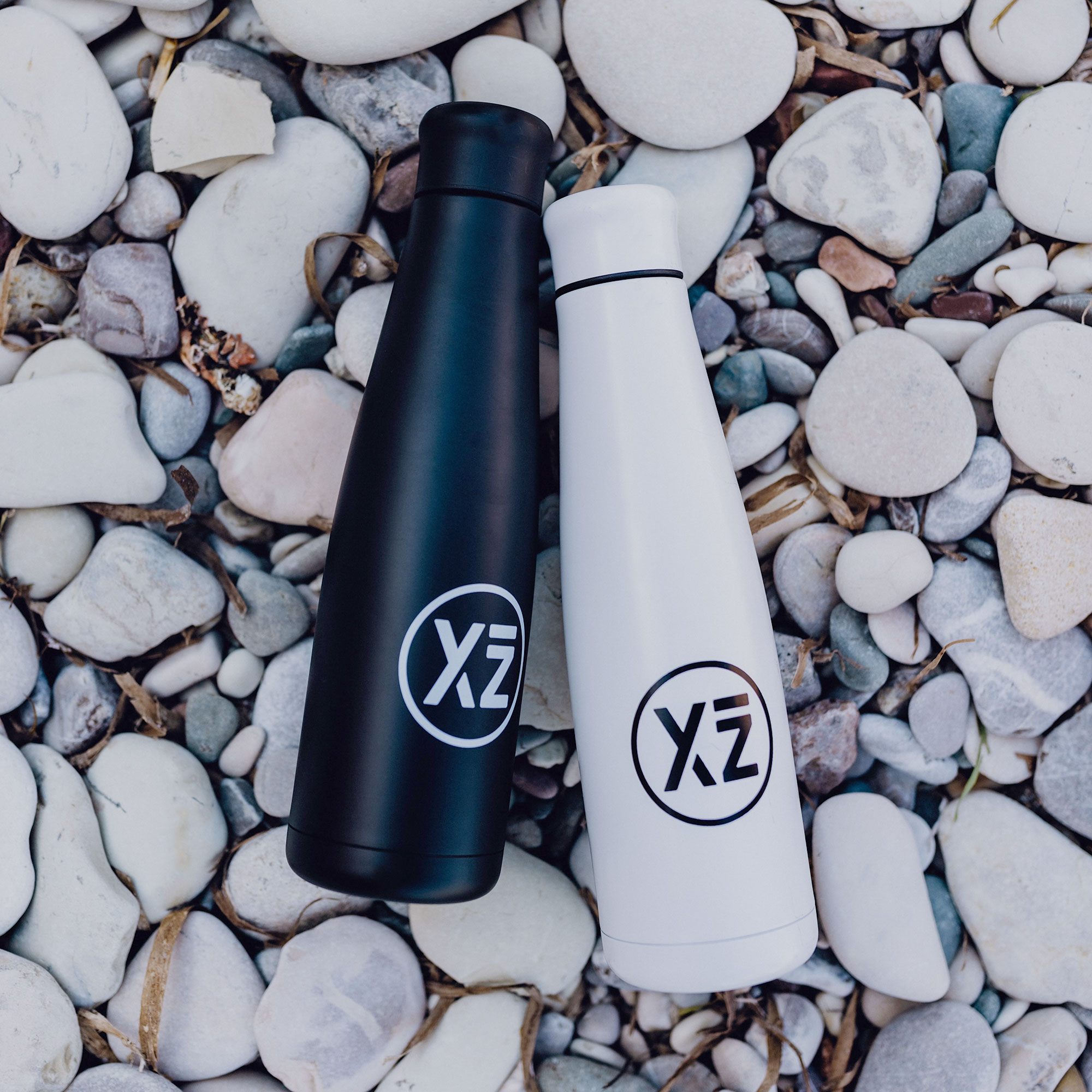 Dos botellas de agua blancas y negras en una playa de guijarros con un logo.
