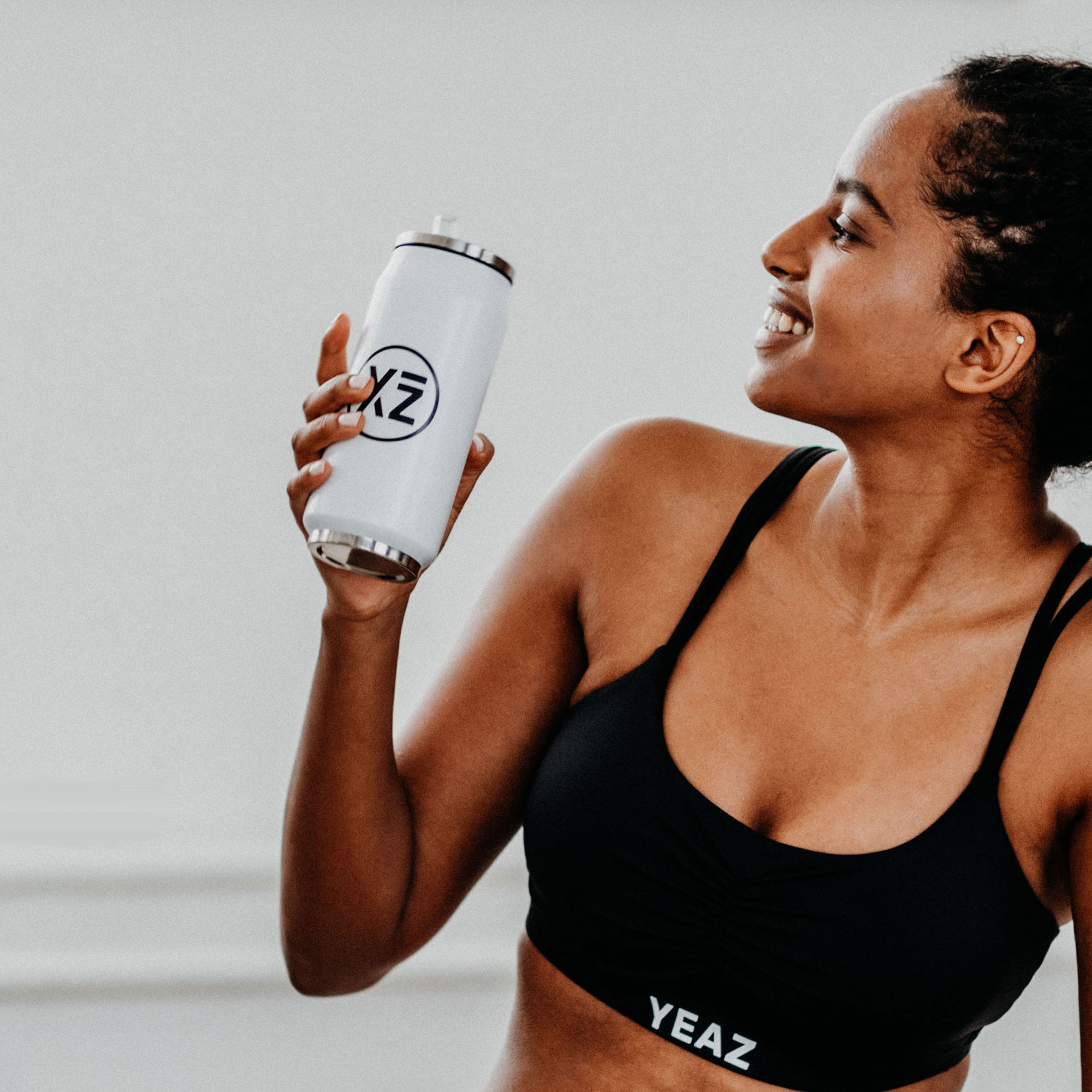 Mujer sosteniendo una botella de agua blanca con un logotipo, sonriendo, con sujetador deportivo negro.