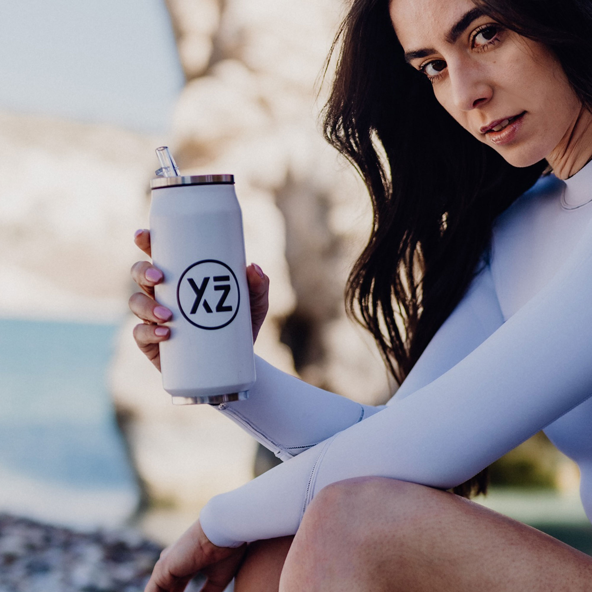 Mujer sosteniendo una botella de agua blanca con logo negro. Lleva un traje de neopreno blanco.
