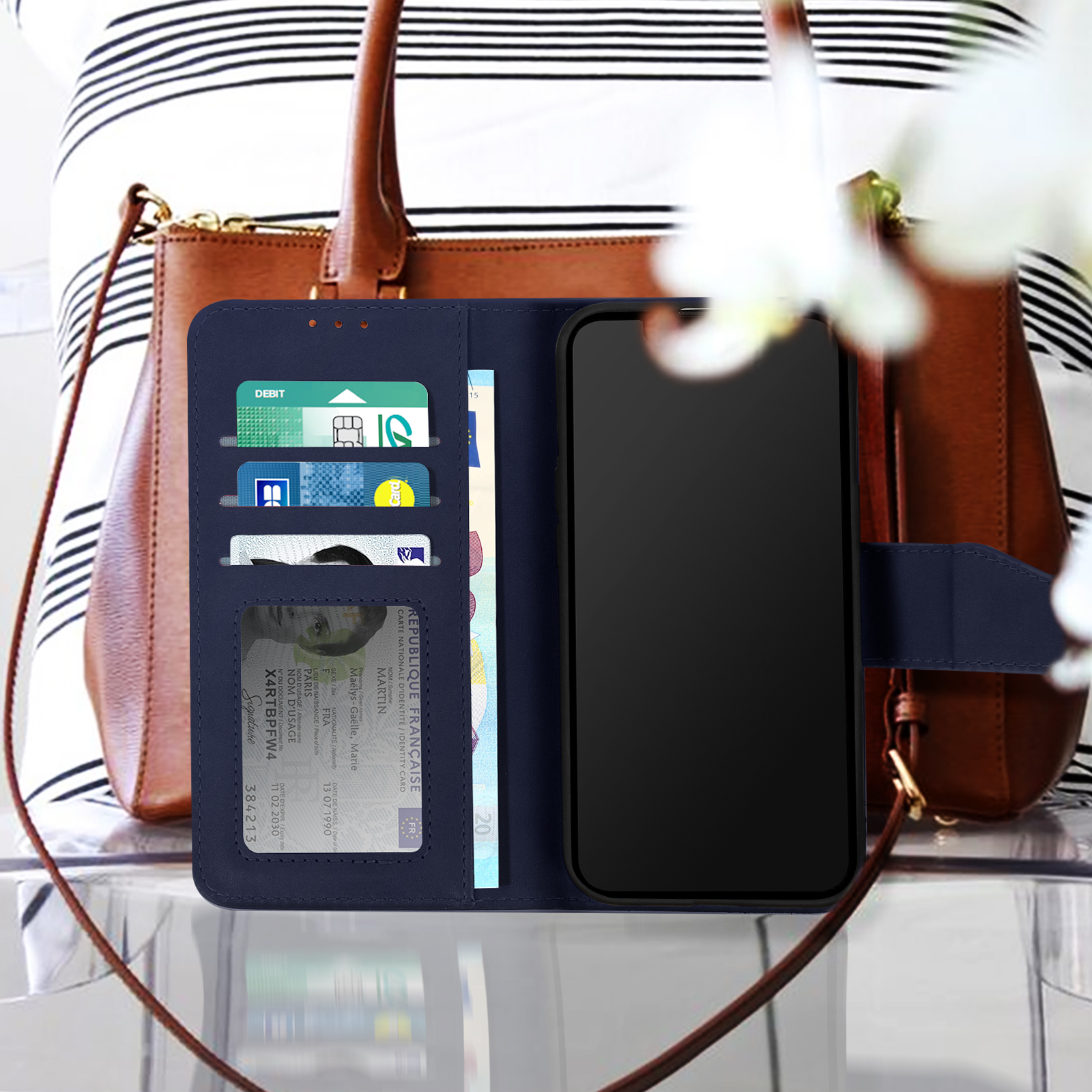 Un téléphone dans un étui portefeuille avec des cartes et une sangle, tenu par une personne, un sac à main marron en arrière-plan.