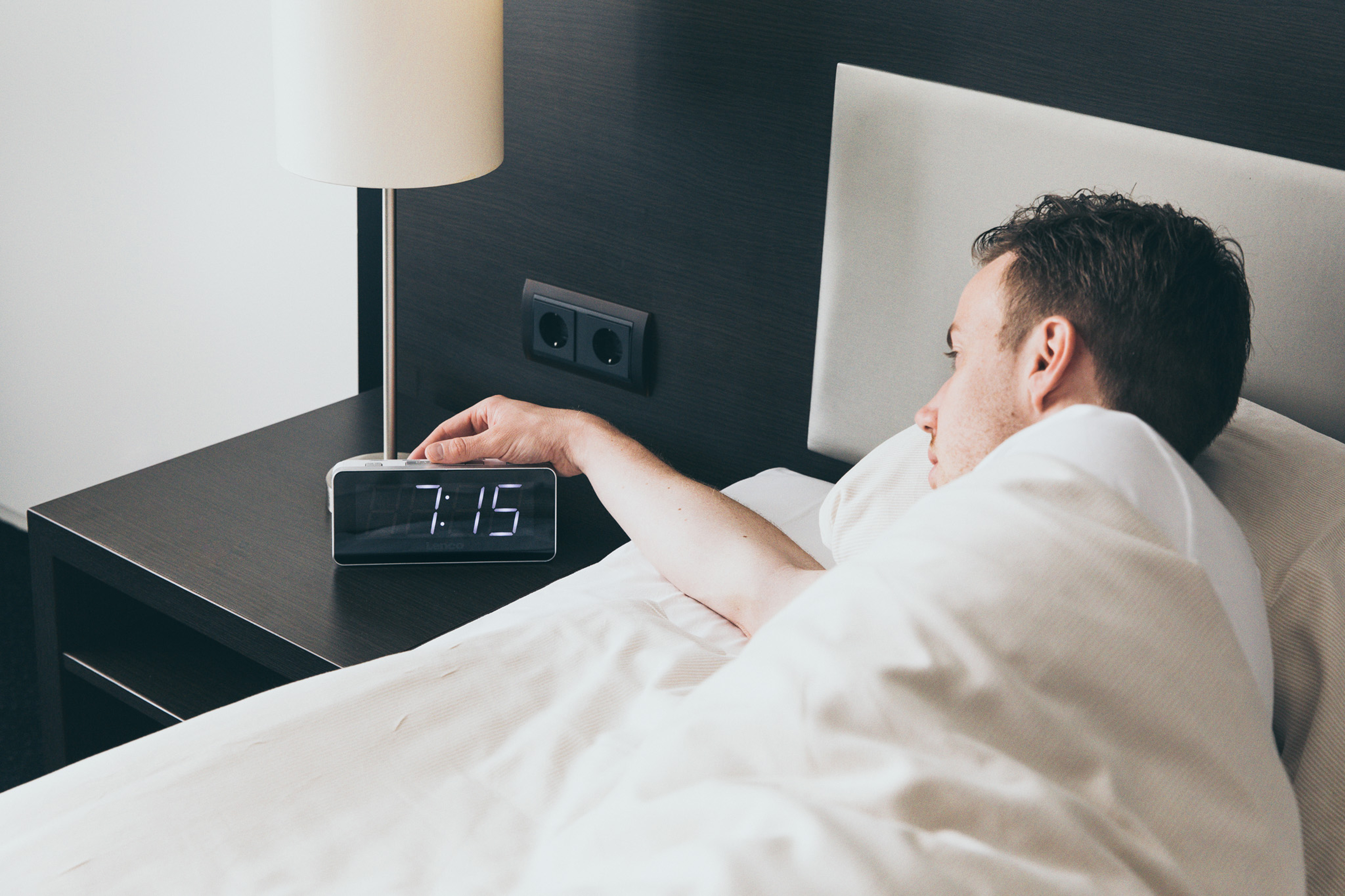 Mężczyzna w łóżku sięgający po cyfrowy zegar pokazujący 7:15, z lampą na stoliku nocnym.